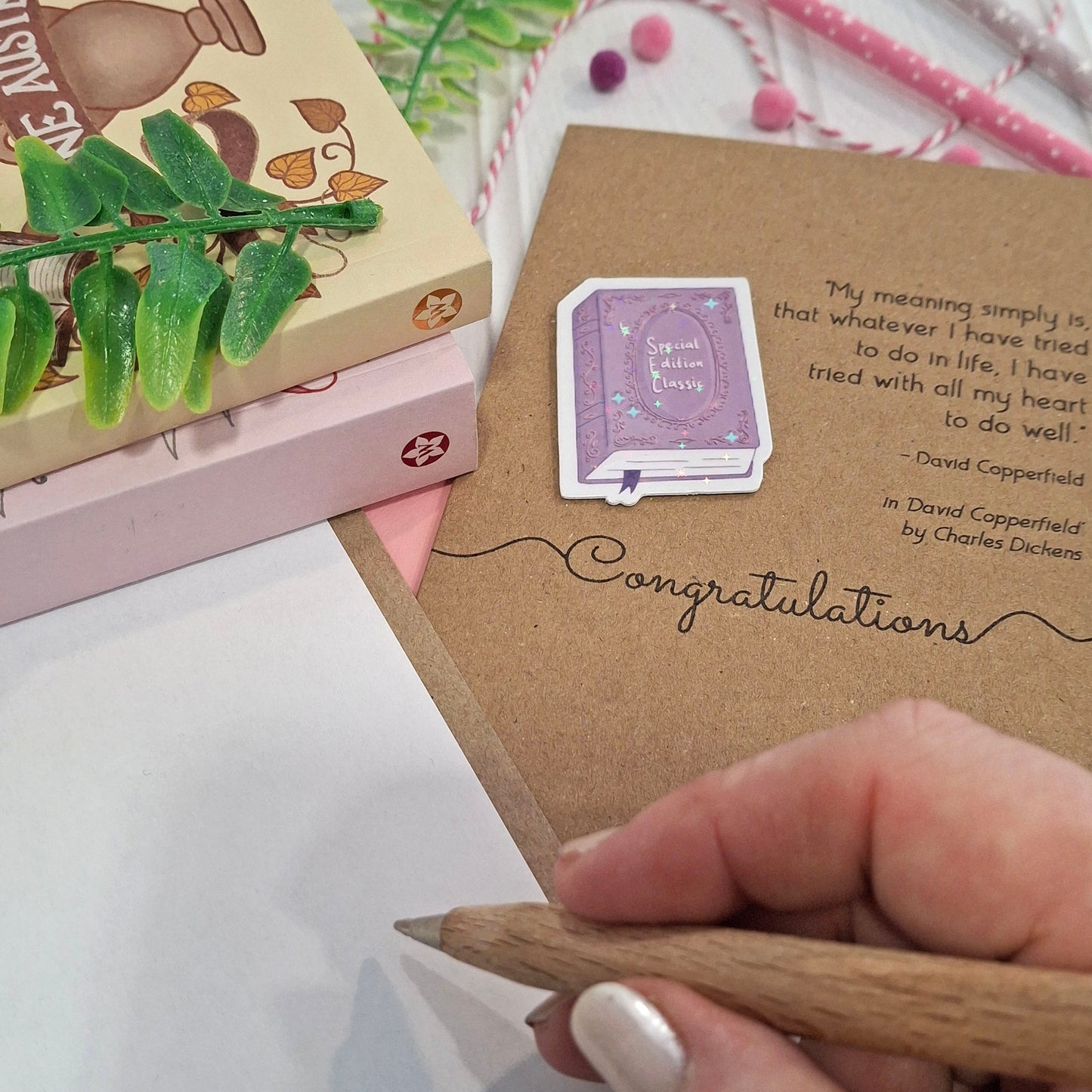 A close-up of a hand holding a wooden pencil writing inside a card. The card features a raised holographic embellishment of a purple special edition classic book, the word Congratulations, and a quote from the book David Copperfield by Charles Dickens.