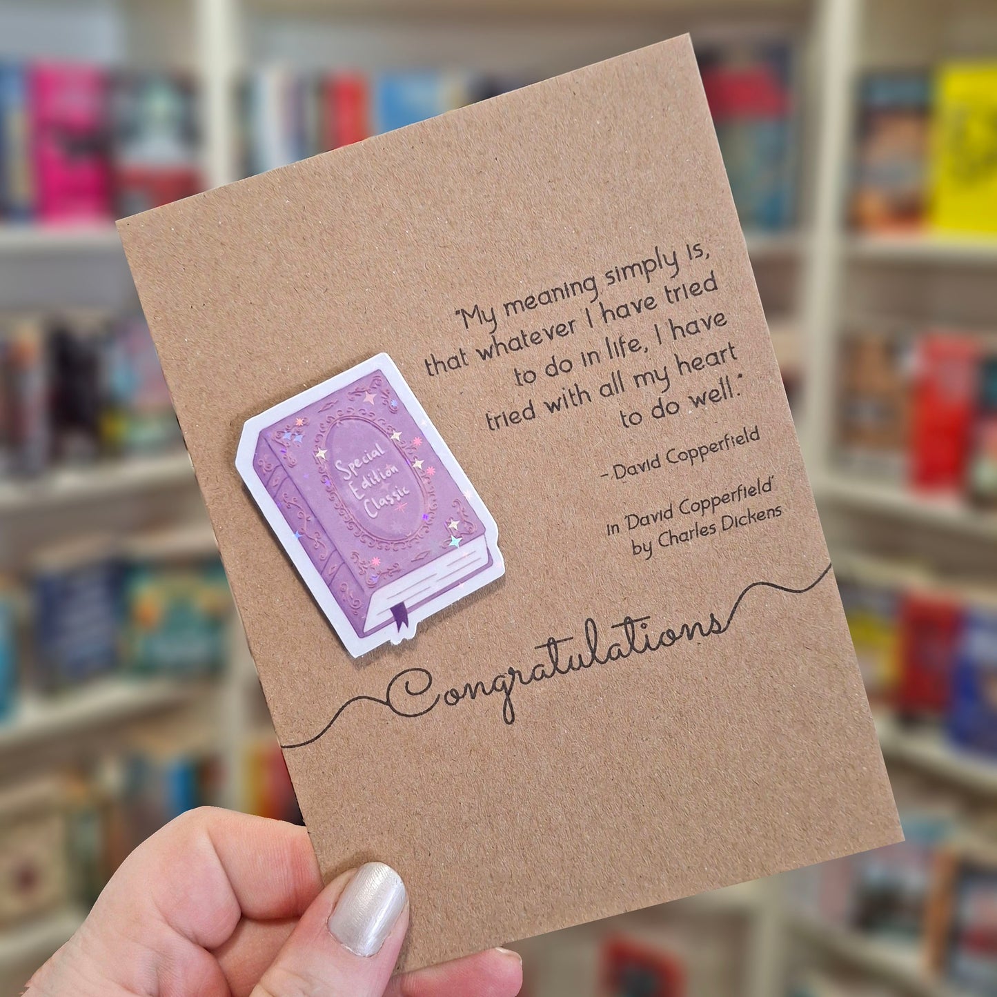 A hand holds a brown kraft congratulations card featuring a raised holographic embellishment of a purple special edition classic book, and a printed quote by Charles Dickens. The background shows a blurred bookshop interior.