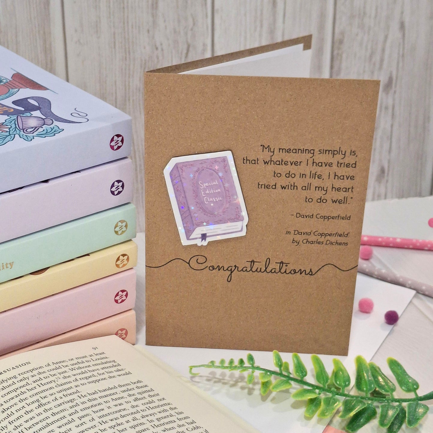 A brown kraft congratulations card styled on a light wood surface with books, an open book, and green leaves. The card features a raised holographic embellishment of a purple special edition classic book, the word Congratulations, and a quote from the book David Copperfield by Charles Dickens.