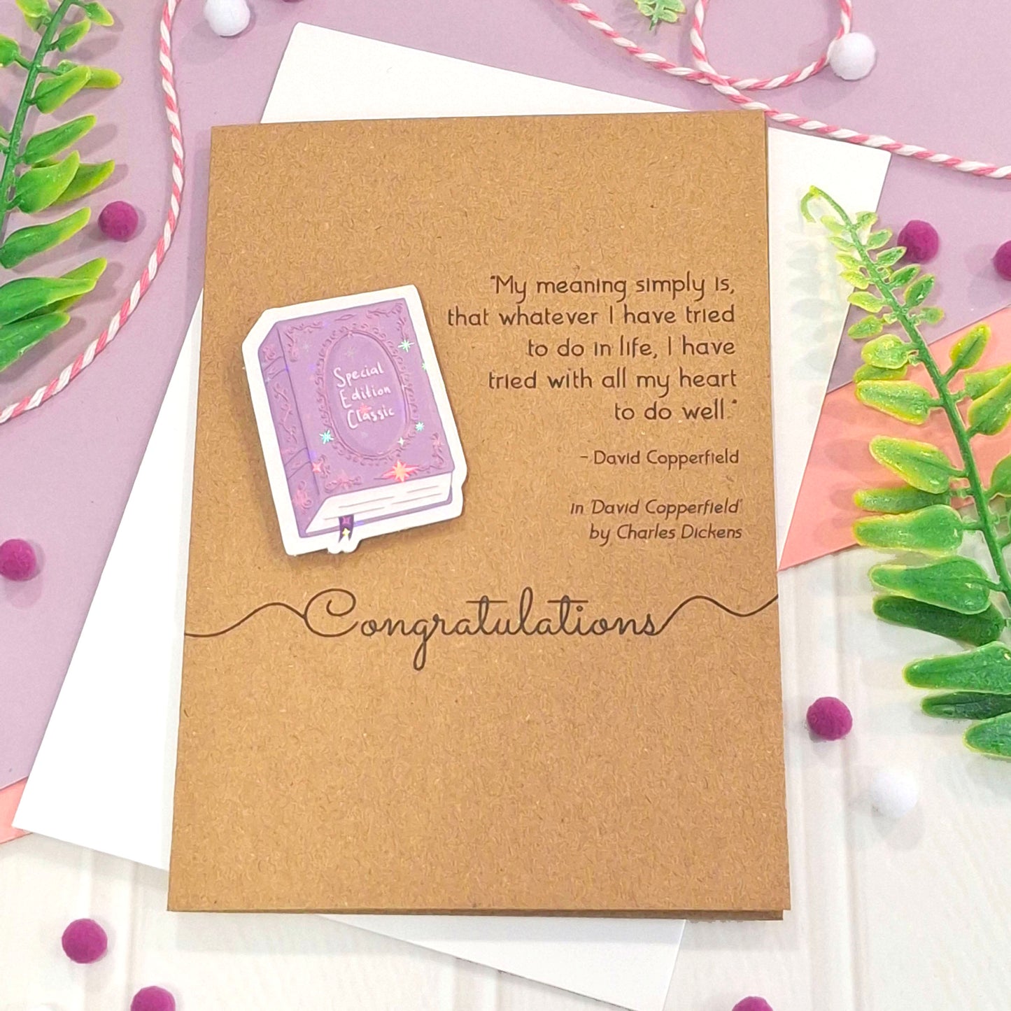 A kraft well done card with a raised, holographic, illustrated story book embellishment in purple, a quote from David Copperfield by Charles Dickens, and the words 'Congratulations'. The card is styled on a white and purple flat-lay background with decorative green leaves, pink and white string, and purple pom-poms.