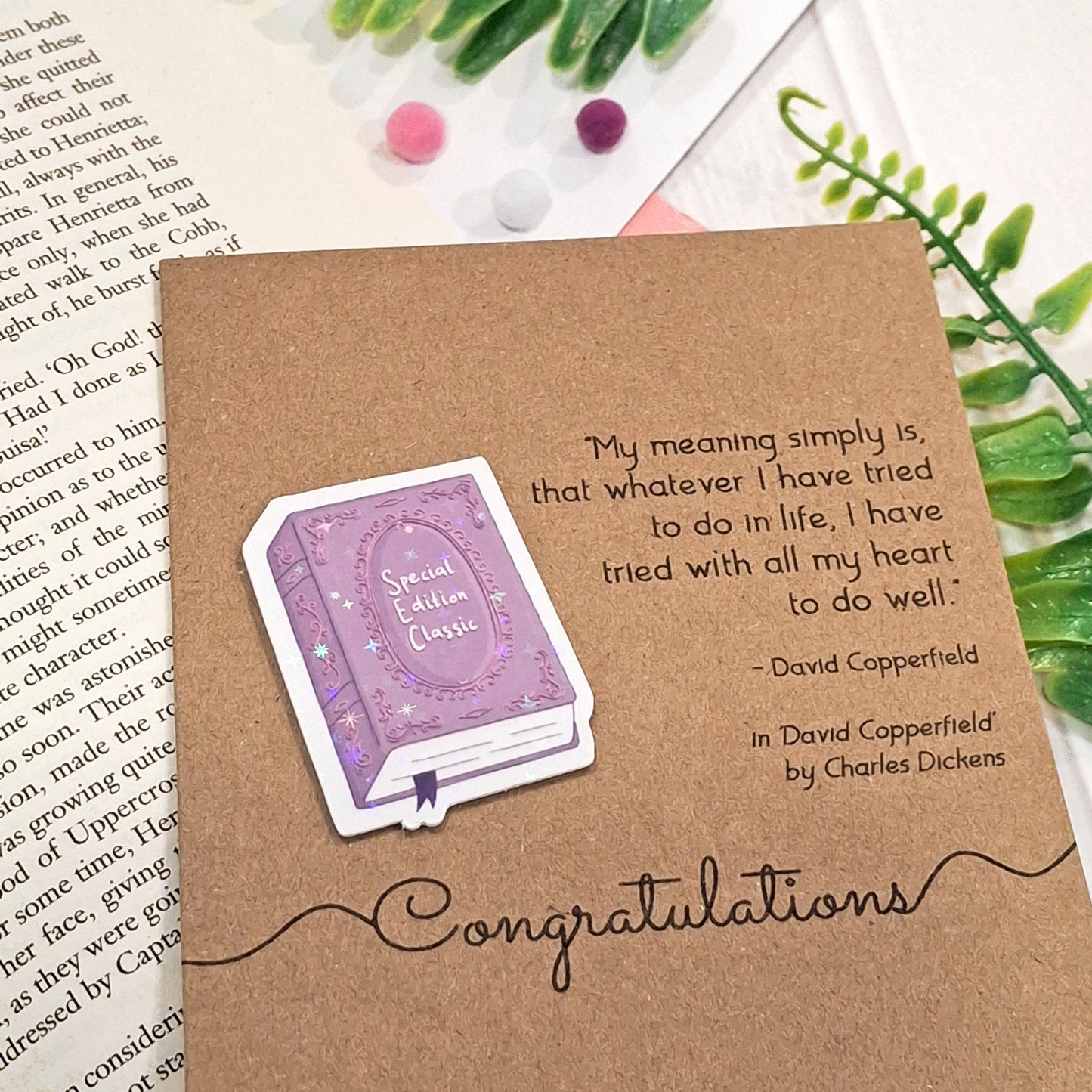 A brown kraft book-themed congratulations card placed on an open book with small decorative pom-poms and green leaves. The card features a raised holographic embellishment of a purple special edition classic book, the word Congratulations, and a quote from the book David Copperfield by Charles Dickens.