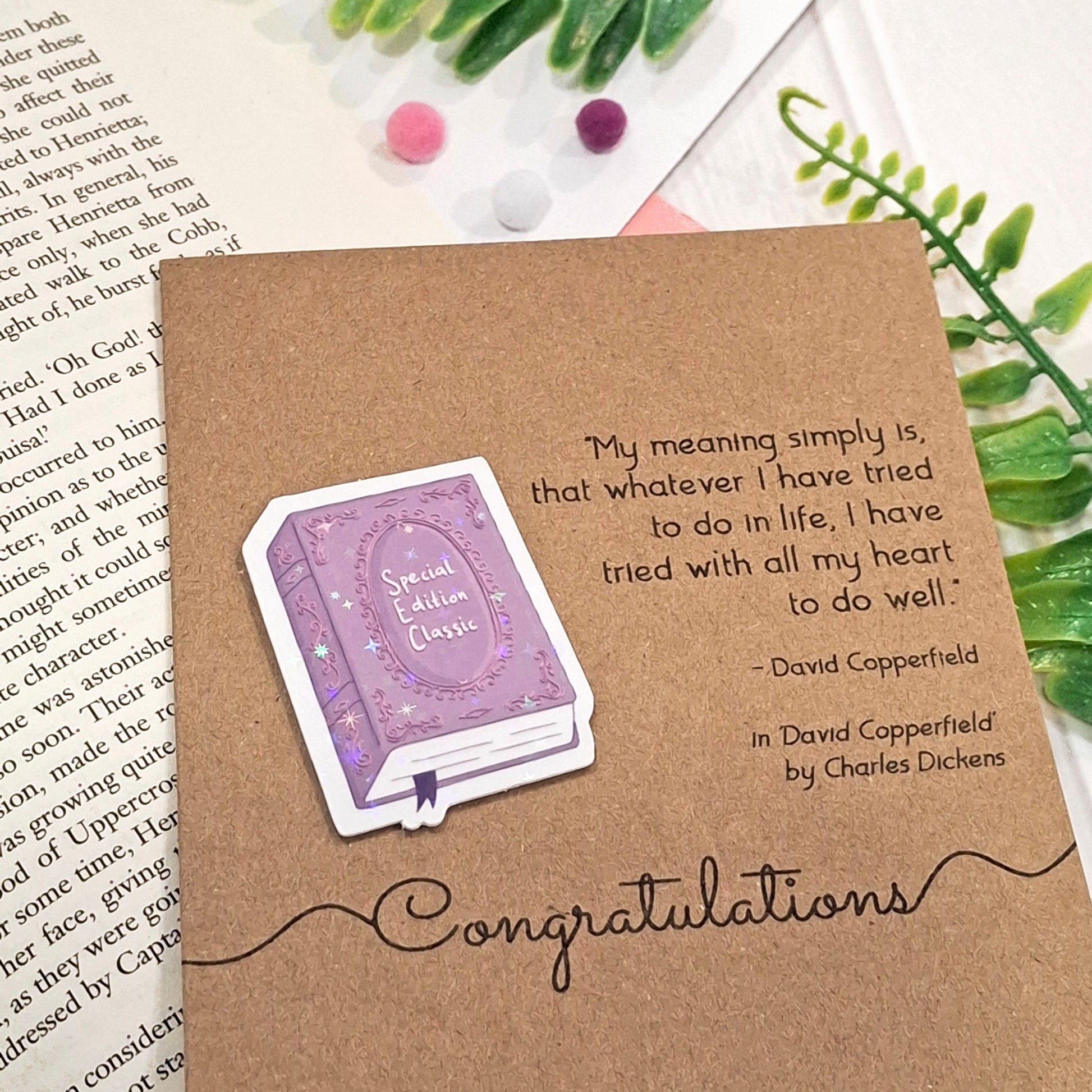 A brown kraft book-themed congratulations card placed on an open book with small decorative pom-poms and green leaves. The card features a raised holographic embellishment of a purple special edition classic book, the word Congratulations, and a quote from the book David Copperfield by Charles Dickens.