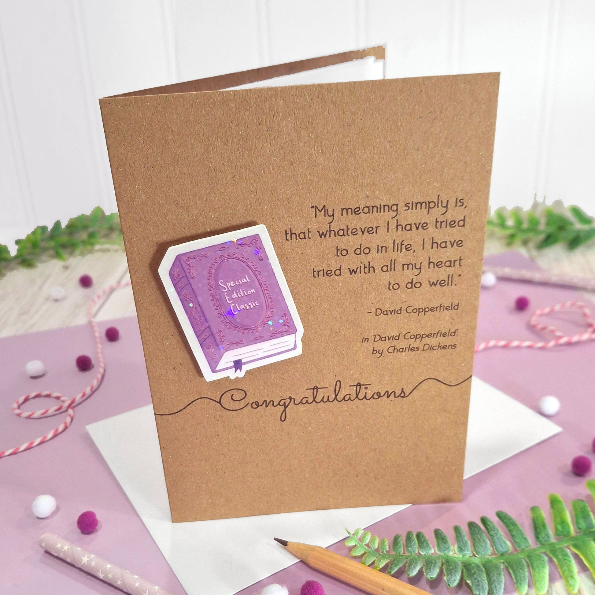 A kraft well done card with a raised, holographic, illustrated story book embellishment in purple, a quote from David Copperfield by Charles Dickens, and the words 'Congratulations', standing on a purple background.