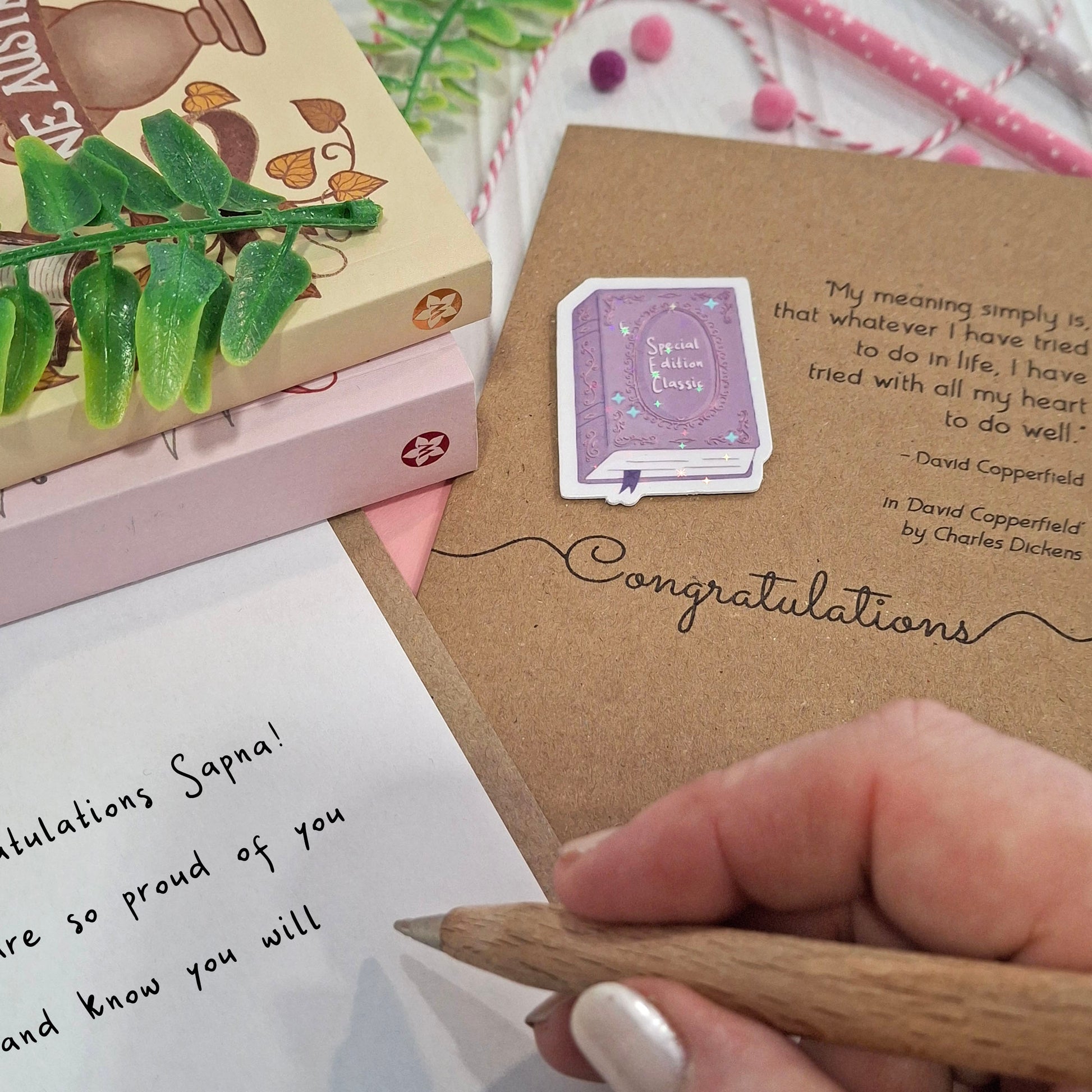 A close-up of a hand holding a wooden pencil writing a well done message inside a card. The card features a raised holographic embellishment of a purple special edition classic book, the word Congratulations, and a quote from the book David Copperfield by Charles Dickens.