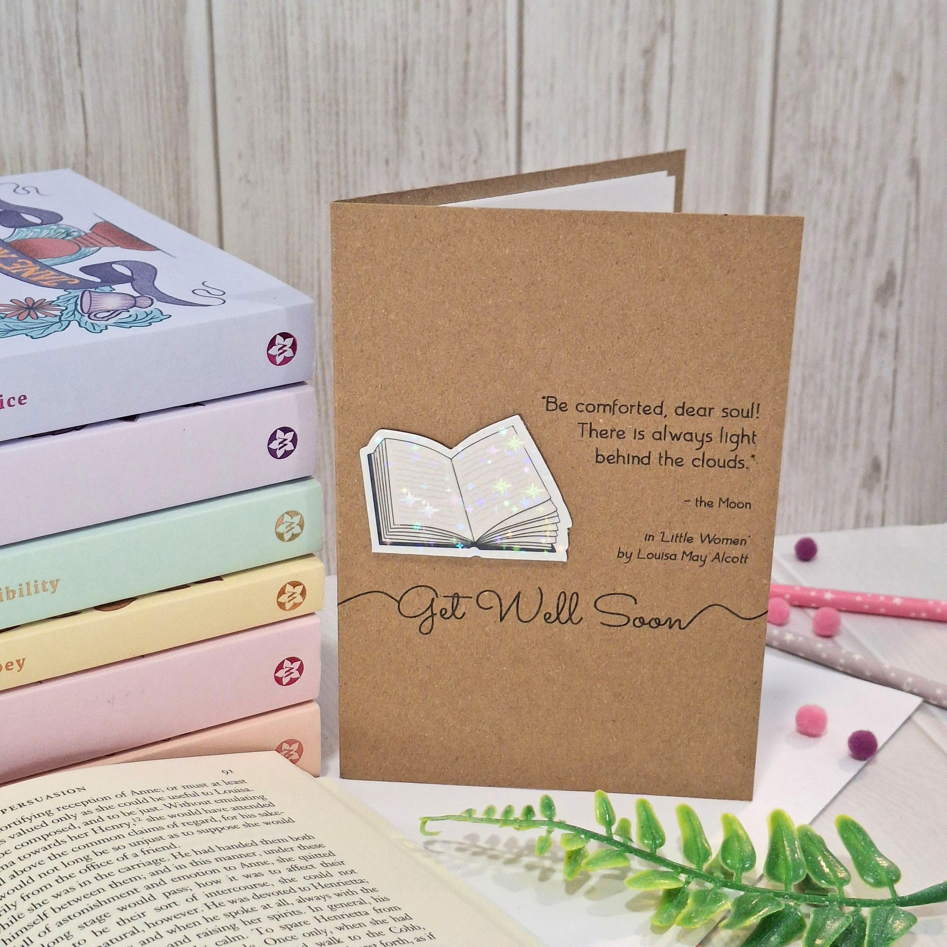 A brown kraft get well card styled with books and a green fern on a light wood background. The card features a raised holographic embellishment of an open book, and reads 'Get well Soon', with a quote from the book Little Women by Louisa May Alcott. 