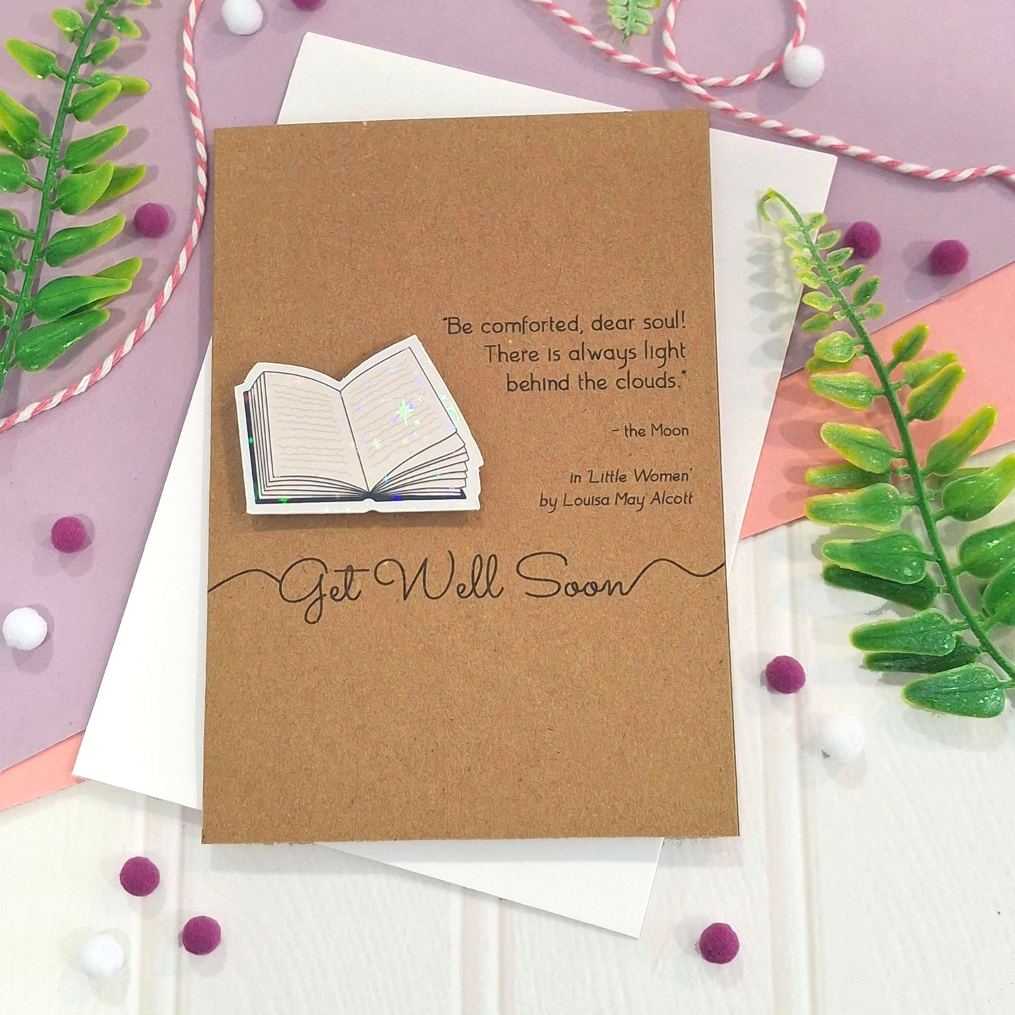 A get well card made from brown kraft card with a raised, holographic, illustrated open book embellishment, a printed quote from the book 'Little Women' by Louisa May Alcott, and the words Get well Soon. The card is styled in a flat-lay arrangement with white envelopes, green leaves, and decorative pom-poms on a pastel purple and white background.