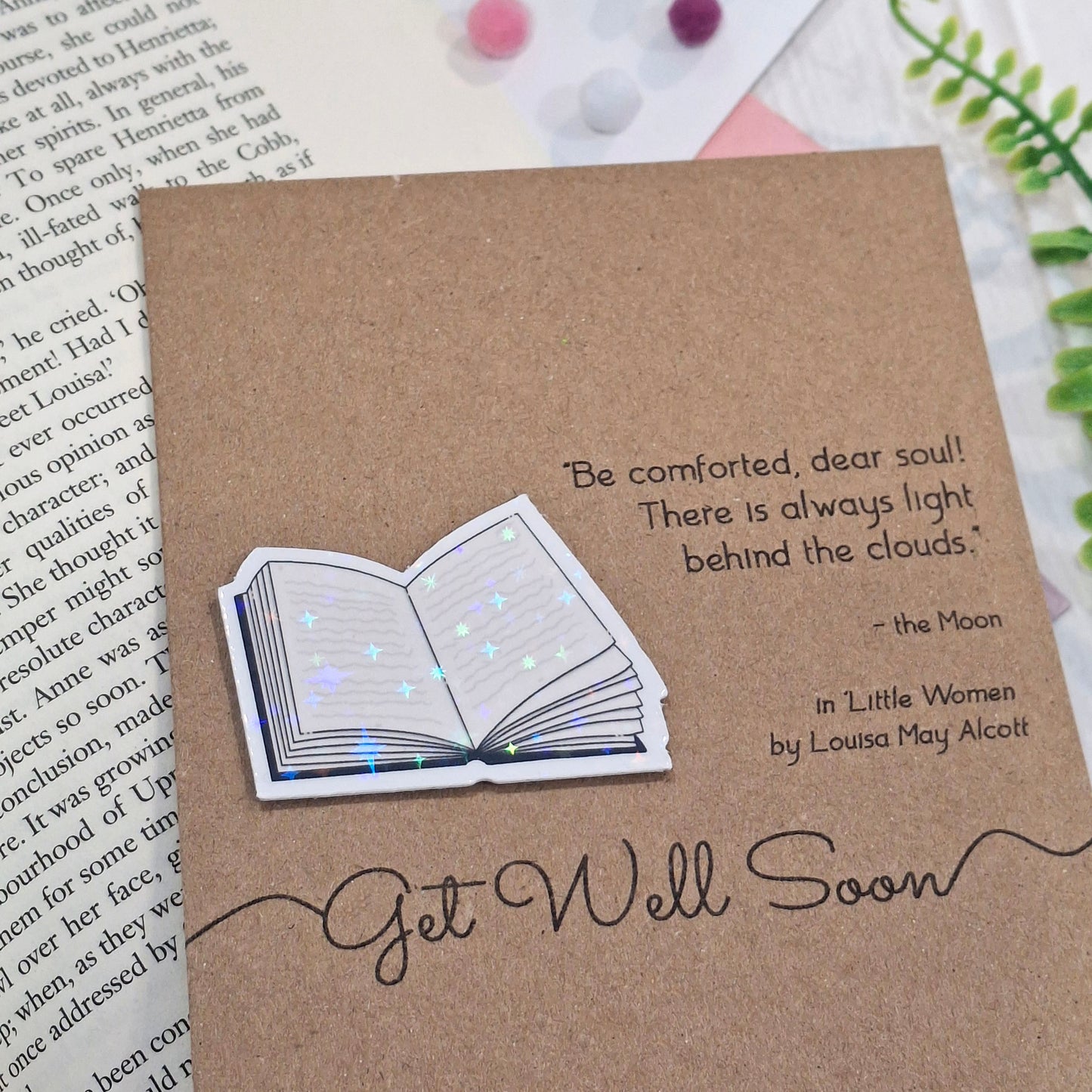 A brown kraft get well soon card styled on an open book page with green ferns in the background. The card features a raised holographic embellishment of an open book, and reads 'Get well Soon', with a quote from the book Little Women by Louisa May Alcott. 
