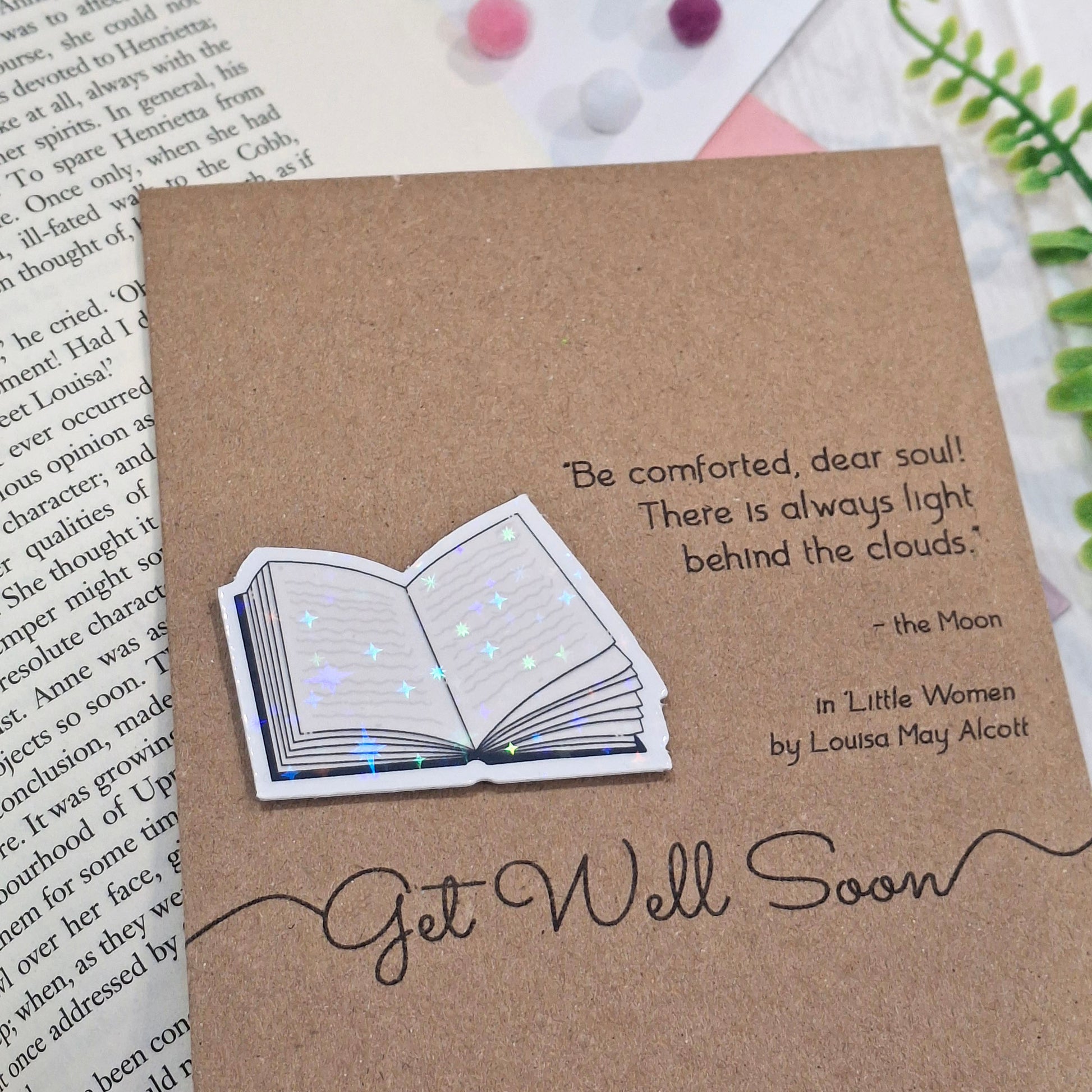 A brown kraft get well soon card styled on an open book page with green ferns in the background. The card features a raised holographic embellishment of an open book, and reads 'Get well Soon', with a quote from the book Little Women by Louisa May Alcott. 