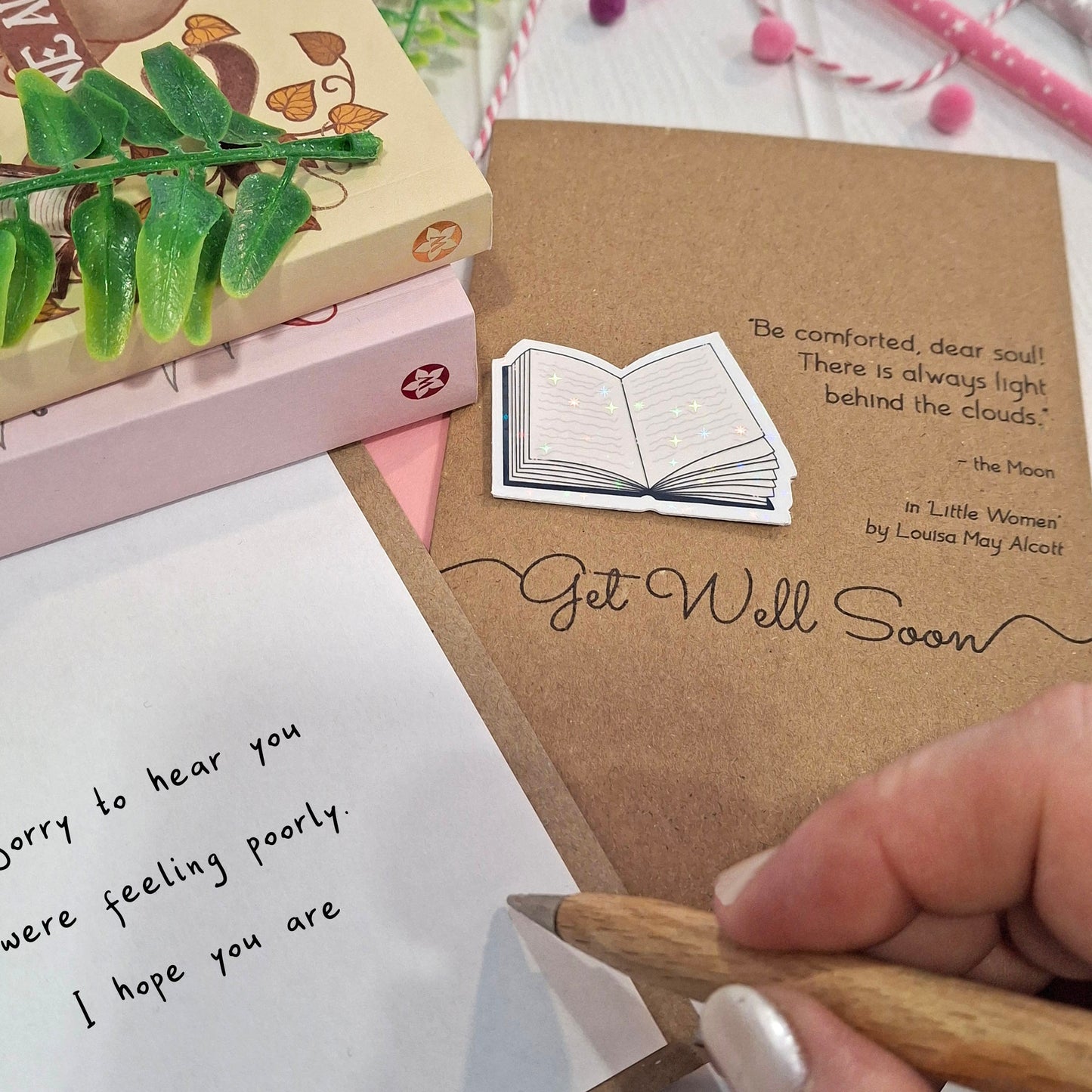 A close-up of a hand holding a wooden pen and writing a get well message  inside a card. The card features a raised holographic embellishment of an open book, and reads 'Get well Soon', with a quote from the book Little Women by Louisa May Alcott. 