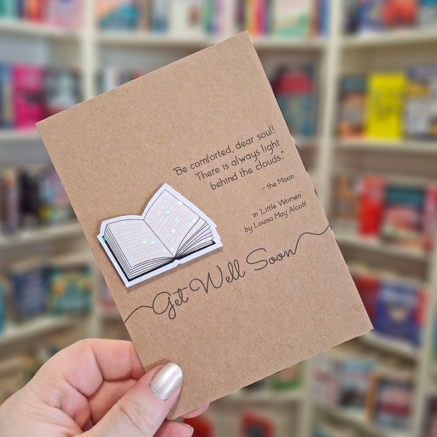 A hand holds a brown kraft get well soon card featuring a raised holographic embellishment of an open book, and a printed quote by Louisa May Alcott. The background shows a blurred bookshop interior.