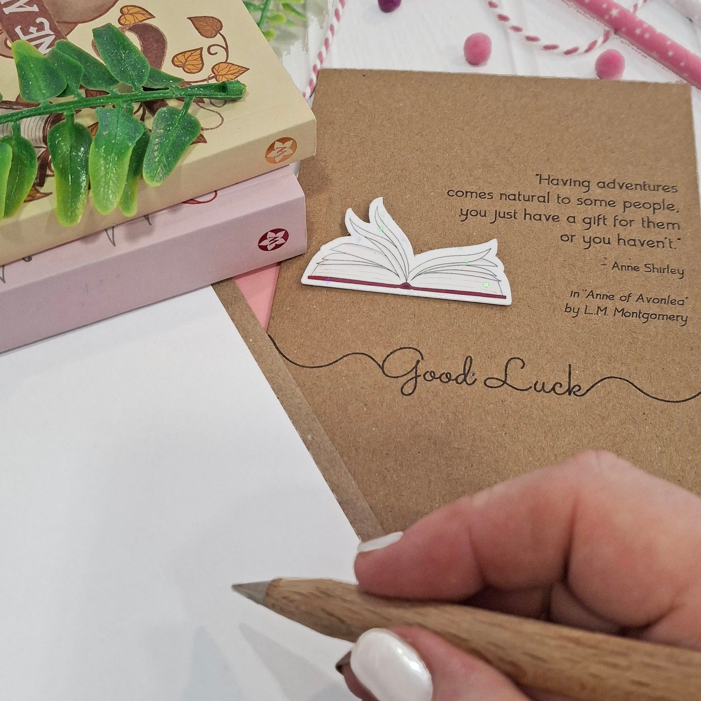 A close up of a hand holding a wooden pen, and writing inside a card. The card features a raised holographic embellishment of an open book with gently fanned pages, the words Good Luck, and a quote from Anne of Avonlea by L.M. Montgomery.