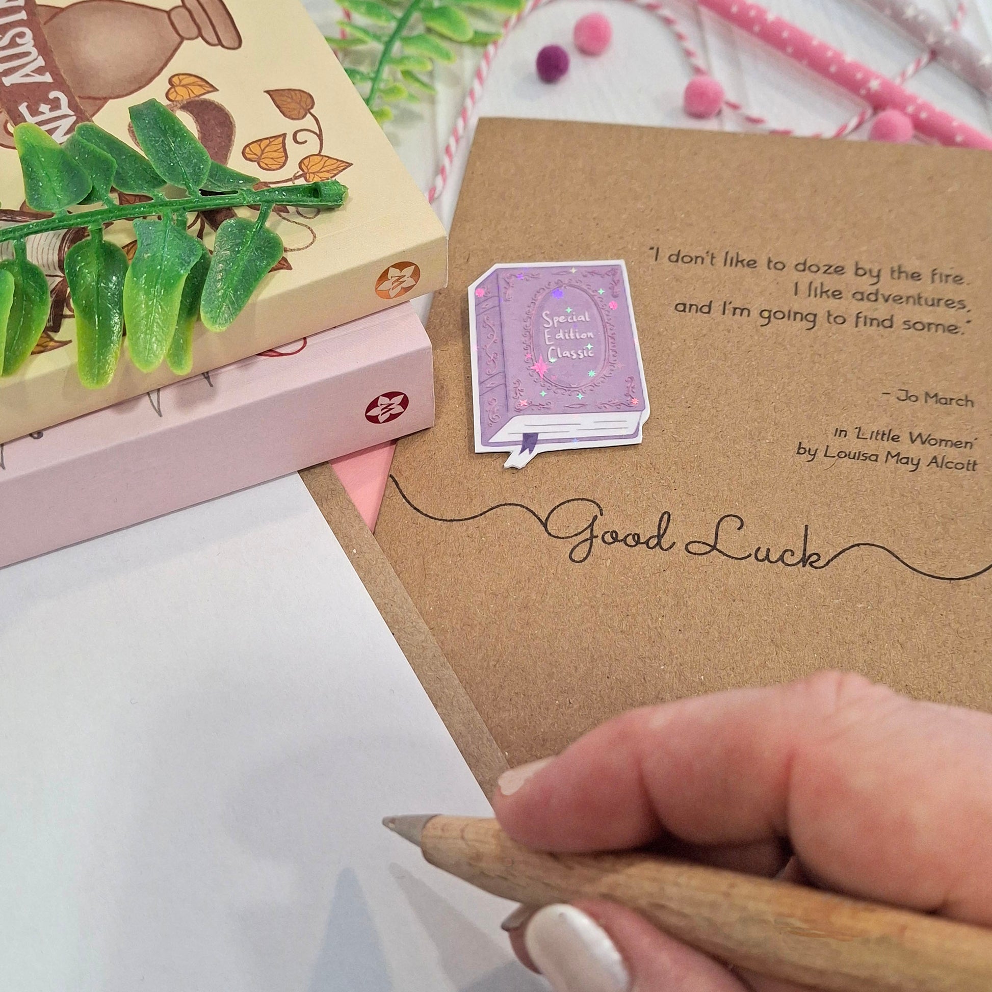 A close-up of a hand holding a wooden pen and writing inside a card. The card features a raised holographic embellishment of a purple special edition classic book, the words Good Luck, and a quote from Little Women by Louisa May Alcott.
