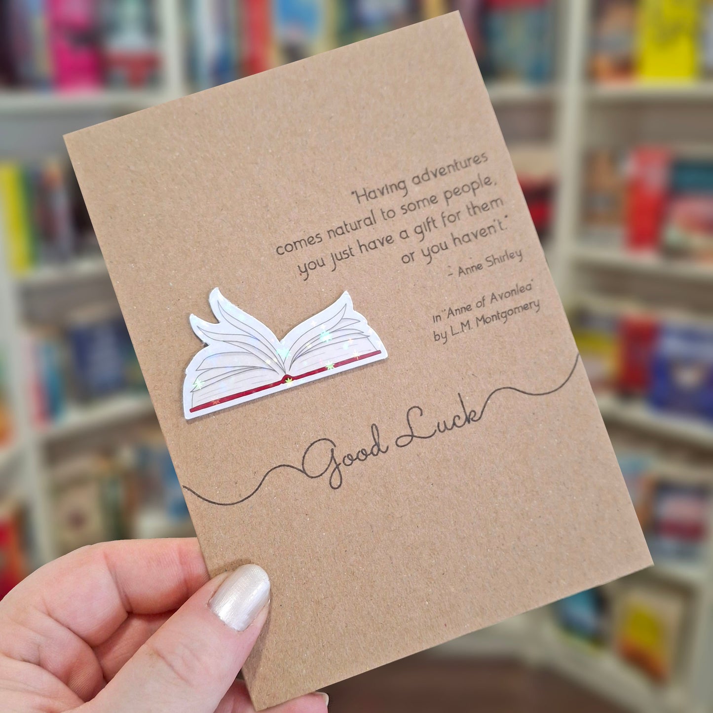 A hand holds a brown kraft good luck card featuring a raised holographic embellishment of an open book with gently fanned pages, and a printed quote by LM Montgomery. The background shows a blurred bookshop interior.