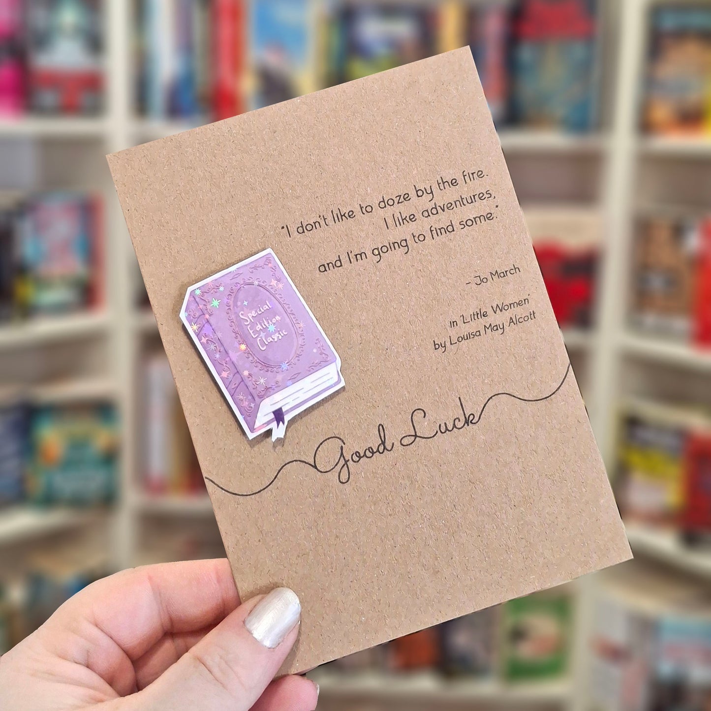 A hand holds a brown kraft good luck card featuring a raised holographic embellishment of a purple special edition classic book, and a printed quote by good luck. The background shows a blurred bookshop interior.
