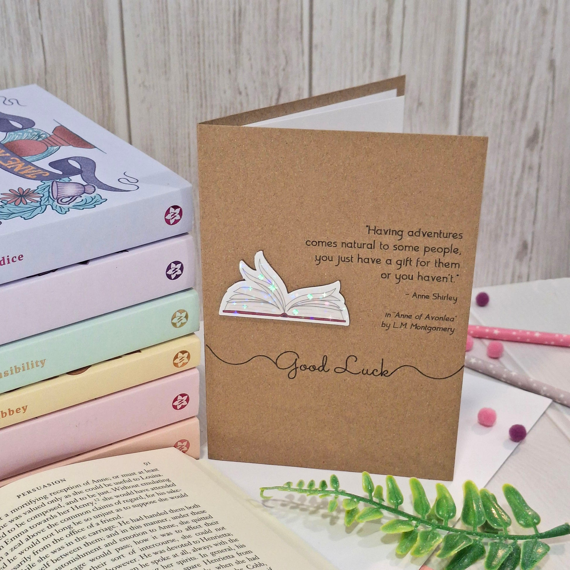 A brown kraft greetings card styled on a tabletop with books, a pencil, and a green sprig. The card features a raised holographic embellishment of an open book with gently fanned pages, the words Good Luck, and a quote from Anne of Avonlea by L.M. Montgomery.