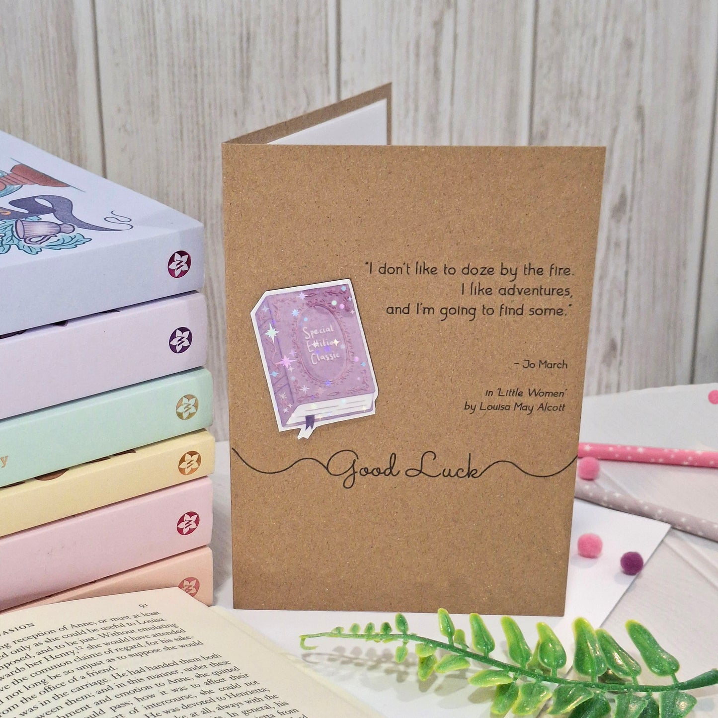 A brown kraft good luck card standing upright. The card features a raised holographic embellishment of a purple special edition classic book, the words Good Luck, and a quote from Little Women by Louisa May Alcott.