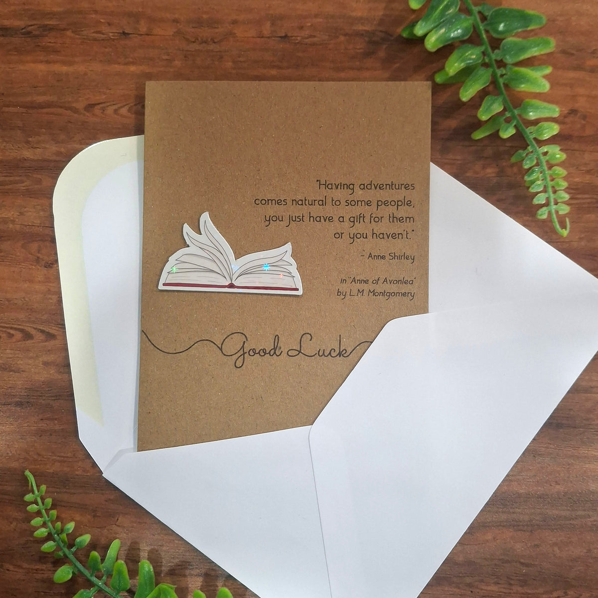 A Good Luck greeting card and white envelope laid on a wooden surface with green leaves all around it. The card is made from brown kraft card and features a raised 3D holographic embellishment of open fluttering book pages, and a quote from Anne of Avonlea by LM Montgomery.