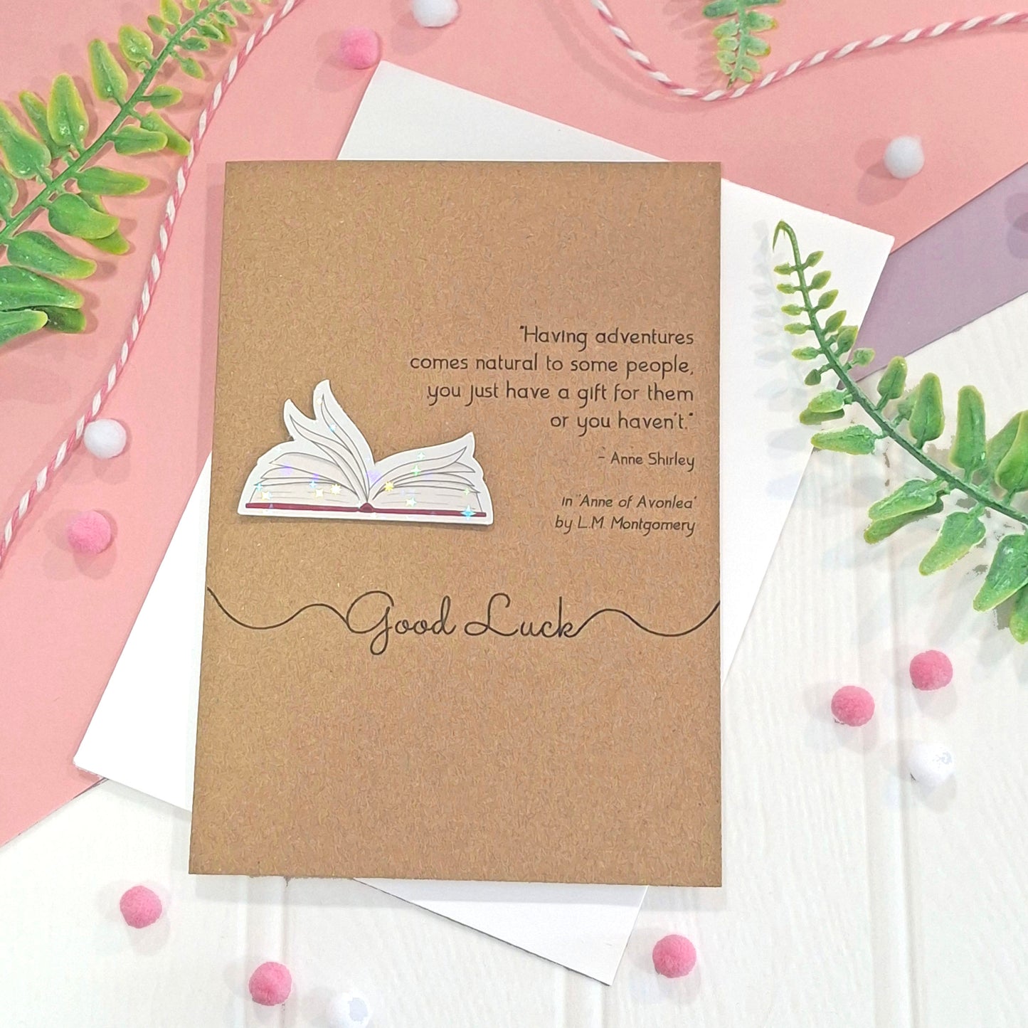 A brown kraft card with illustrated book pages in a raised, holographic embellishment, the words 'Good Luck' and a quote from Anne of Avonlea by LM Montgomery, styled on a pink and white surface with green leaves and pom-poms.