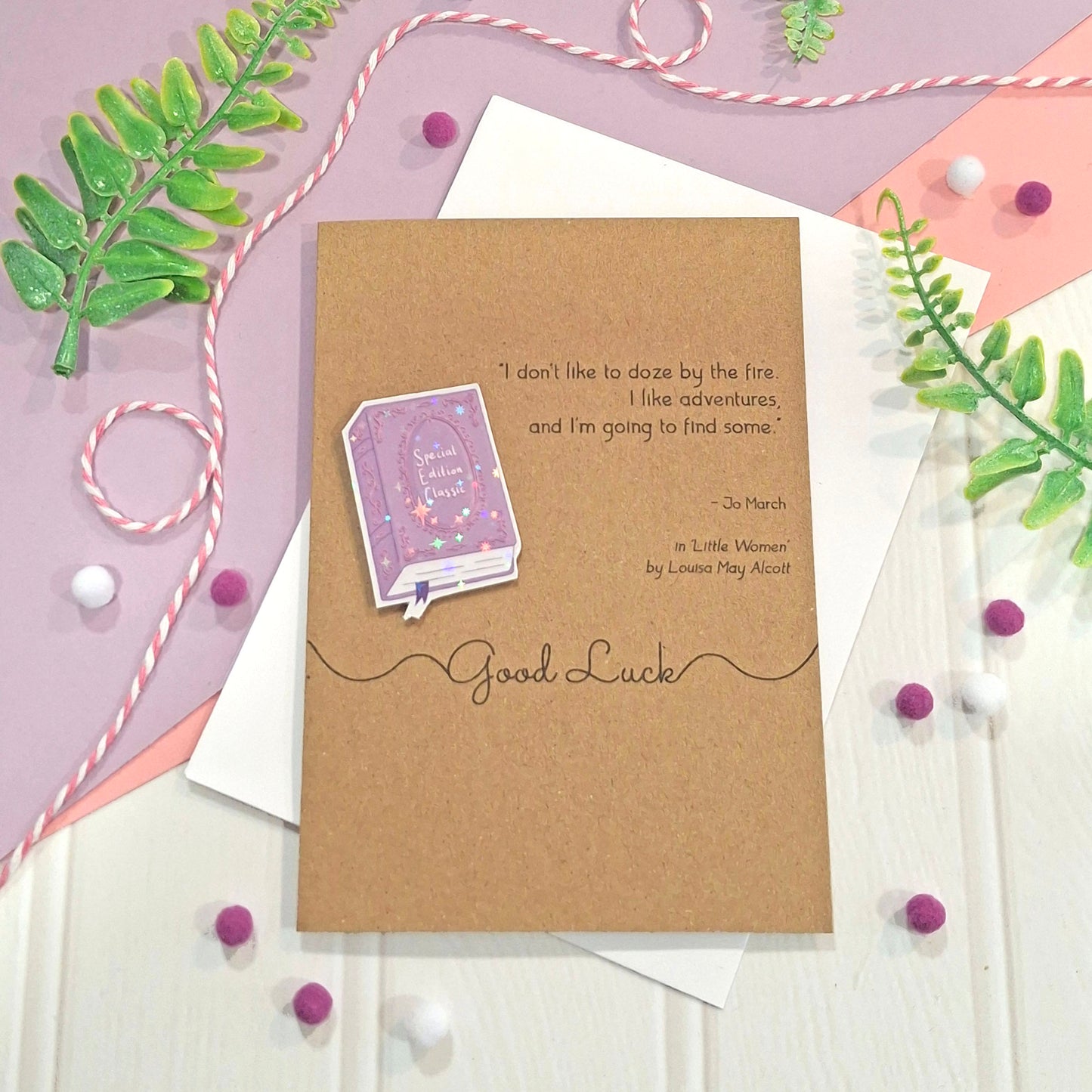 A brown kraft card with a raised, holographic, illustrated story book embellishment in purple, and a quote by Louisa May Alcott from the book Little Women. The card reads Good Luck, and is styled on a white wood surface with envelopes, green leaves, and small pom-poms.
