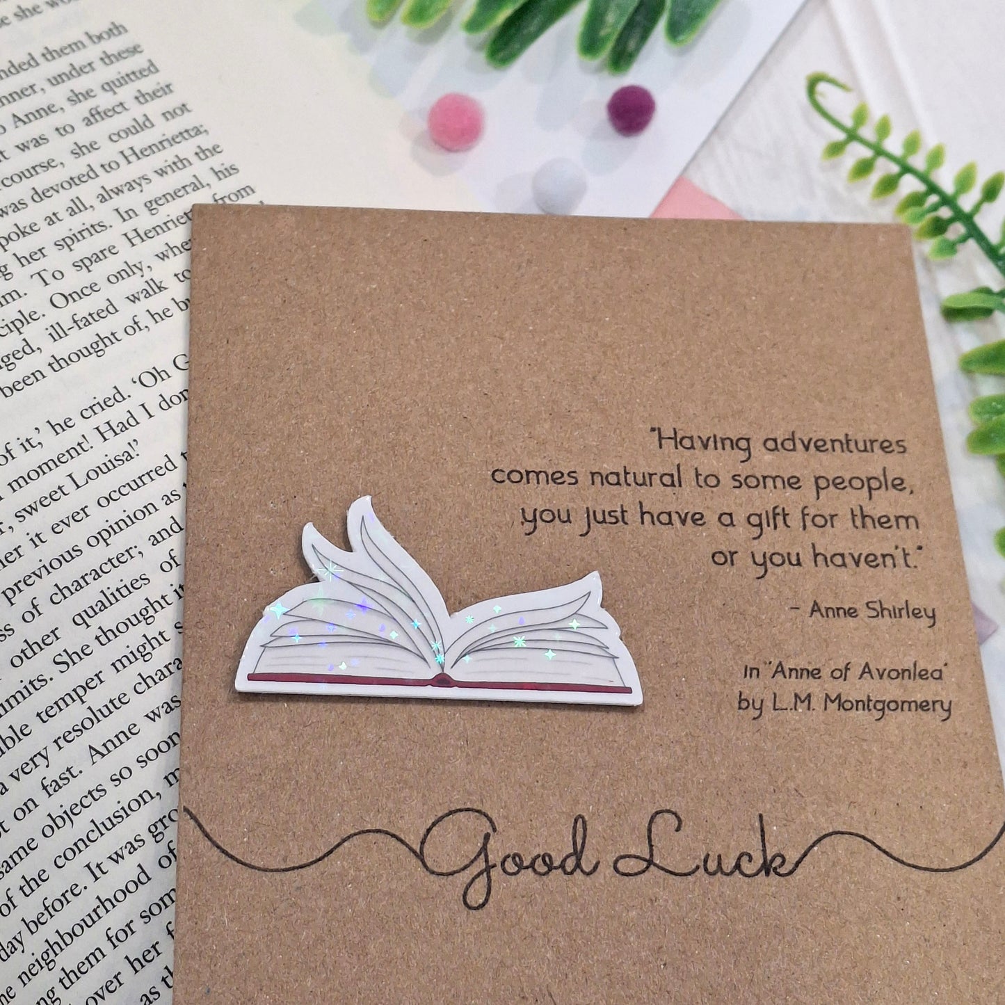 A brown kraft card styled on top of an open book with decorative green leaves and small pom-poms. The card features a raised holographic embellishment of an open book with gently fanned pages, the words Good Luck, and a quote from Anne of Avonlea by L.M. Montgomery.