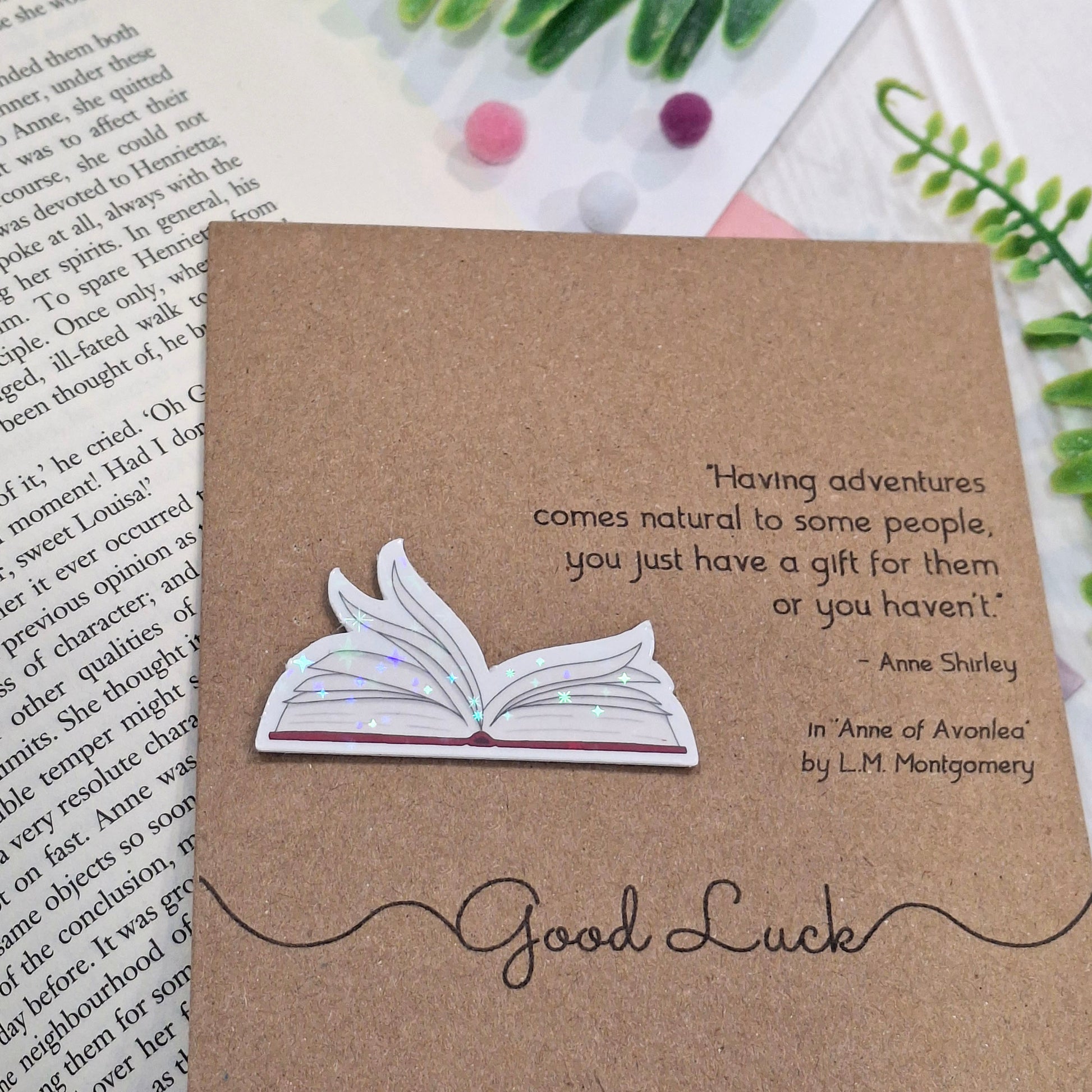 A brown kraft card styled on top of an open book with decorative green leaves and small pom-poms. The card features a raised holographic embellishment of an open book with gently fanned pages, the words Good Luck, and a quote from Anne of Avonlea by L.M. Montgomery.