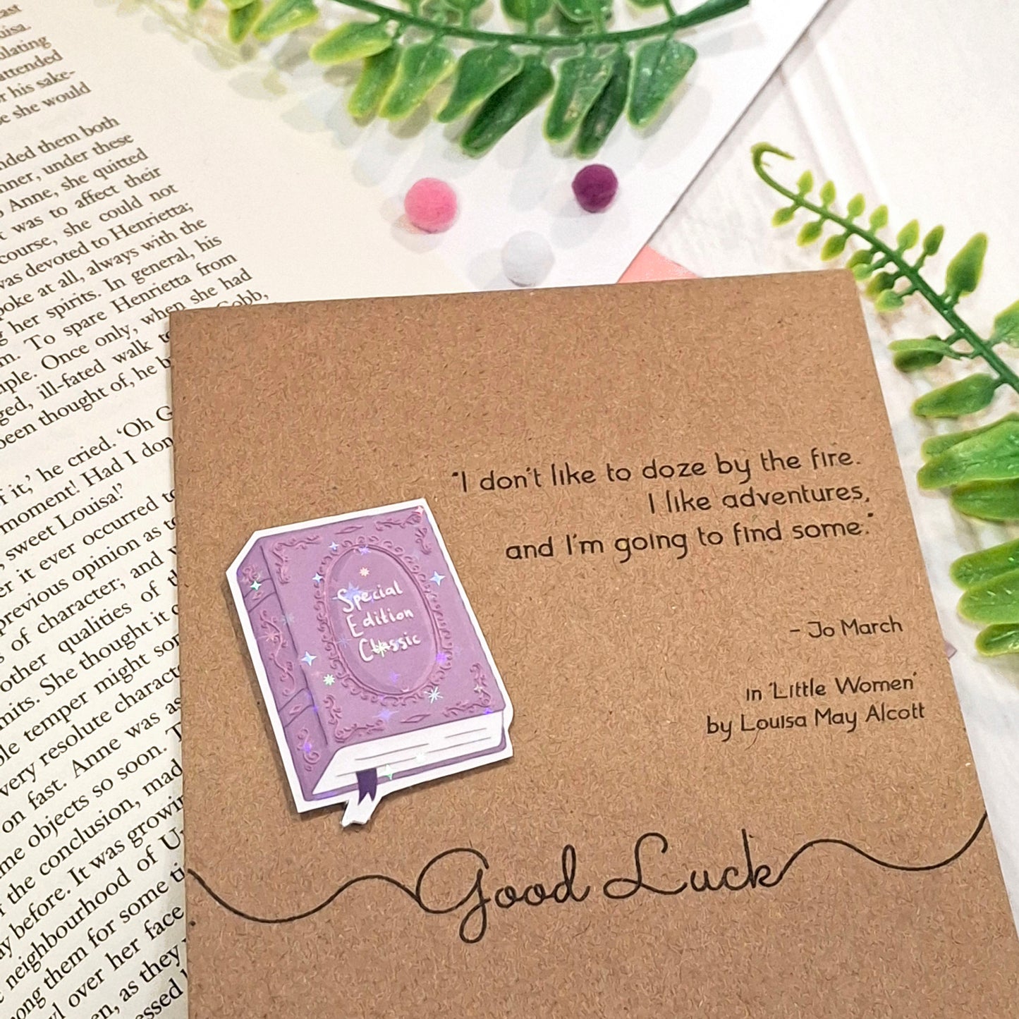 A brown kraft card styled on top of an open book with green leaves and small pom-poms. The card features a raised holographic embellishment of a purple special edition classic book, the words Good Luck, and a quote from Little Women by Louisa May Alcott.