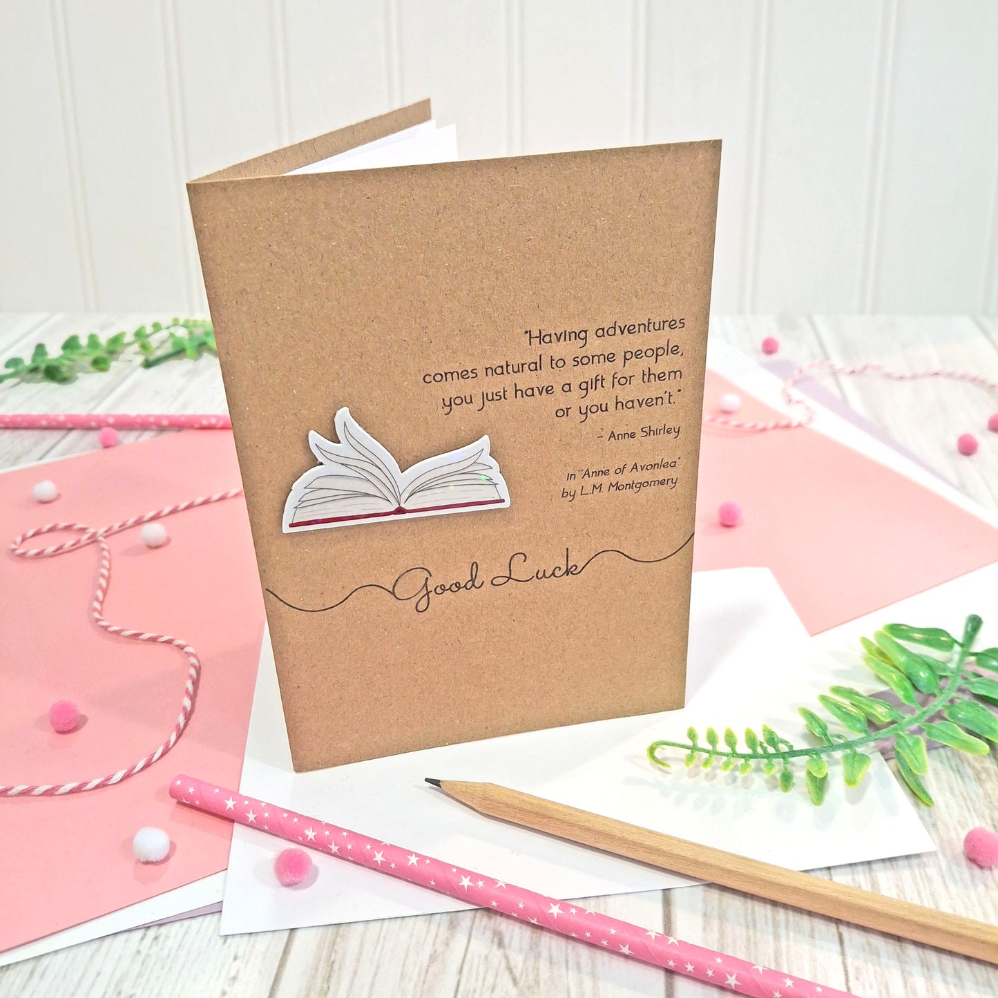 A brown kraft card that reads 'good luck', and features illustrated book pages in a raised, holographic embellishment, and a quote by LM Montgomery from the book Anne of Avonlea styled on a light wood tabletop with pink paper, pencils, and greenery.