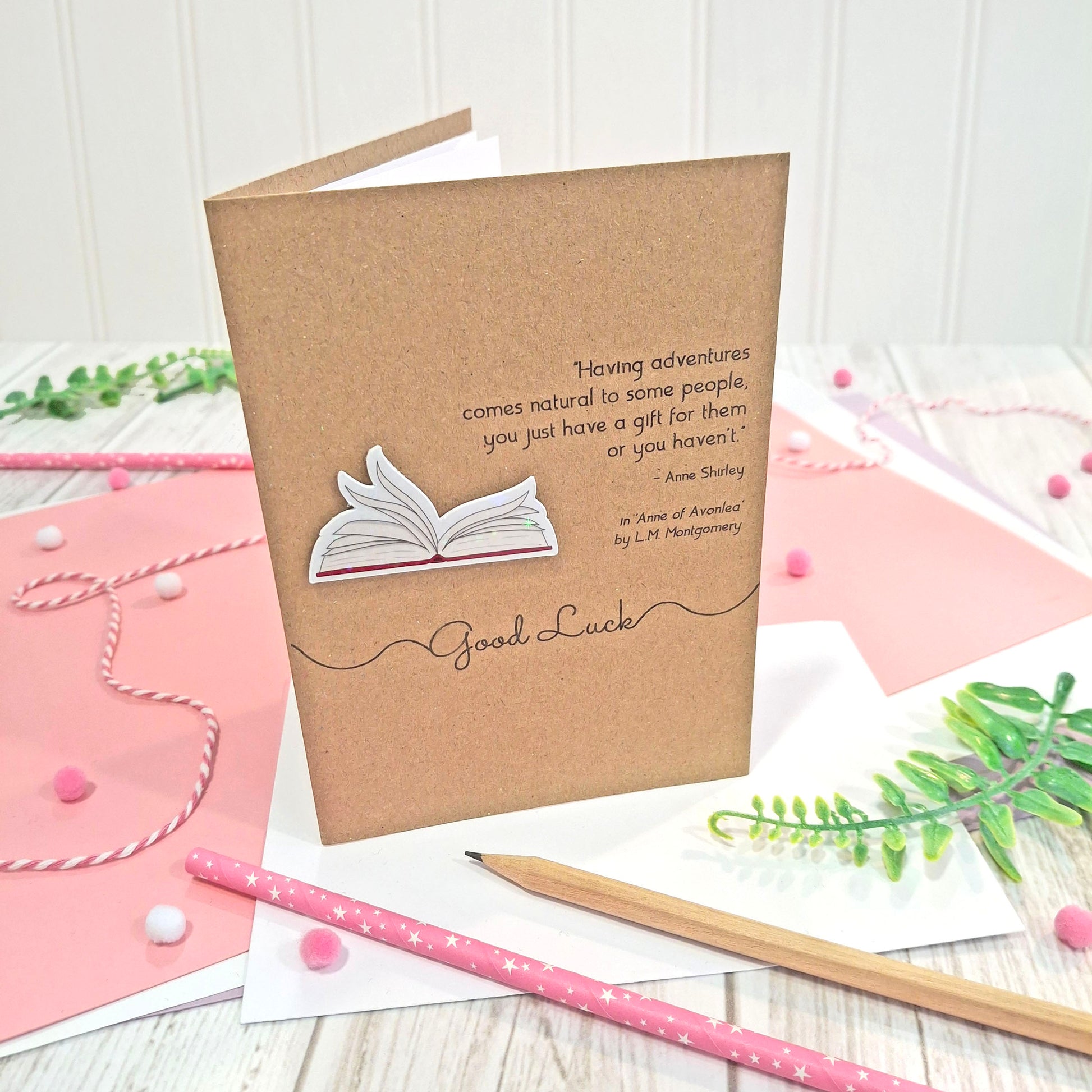 A brown kraft card that reads 'good luck', and features illustrated book pages in a raised, holographic embellishment, and a quote by LM Montgomery from the book Anne of Avonlea styled on a light wood tabletop with pink paper, pencils, and greenery.