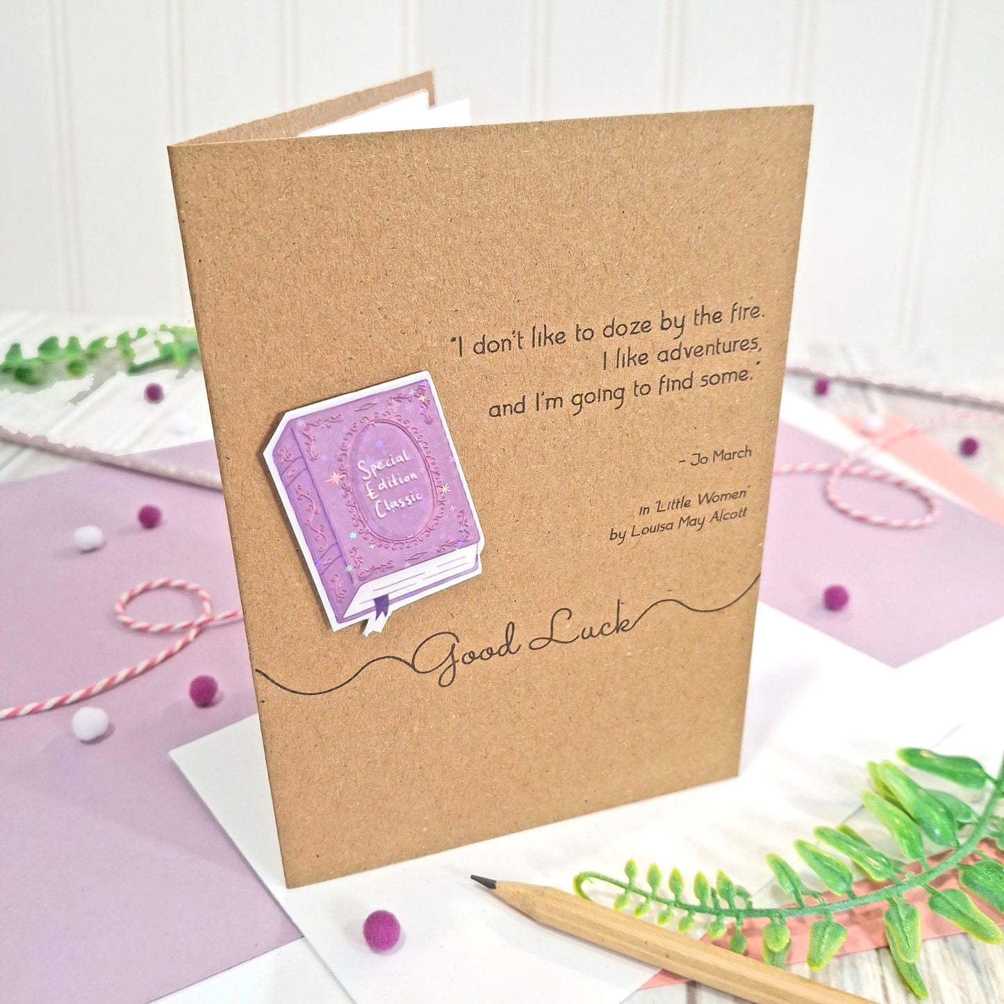A handmade greeting card made from brown kraft card with a raised, holographic, illustrated story book embellishment in purple, a quote from 'Little Women', by Louisa May Alcott, and the words 'Good Luck' all displayed on a pastel purple desk with craft props.