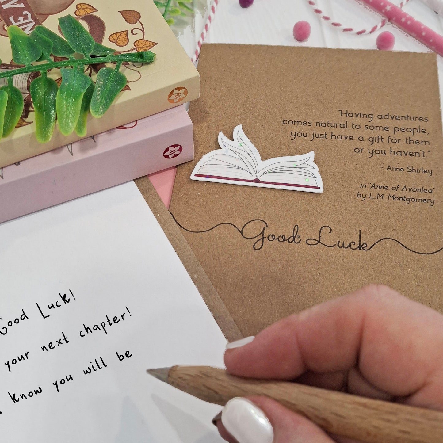 A close up of a hand holding a wooden pen, and writing a good luck message inside a card. The card features a raised holographic embellishment of an open book with gently fanned pages, the words Good Luck, and a quote from Anne of Avonlea by L.M. Montgomery.