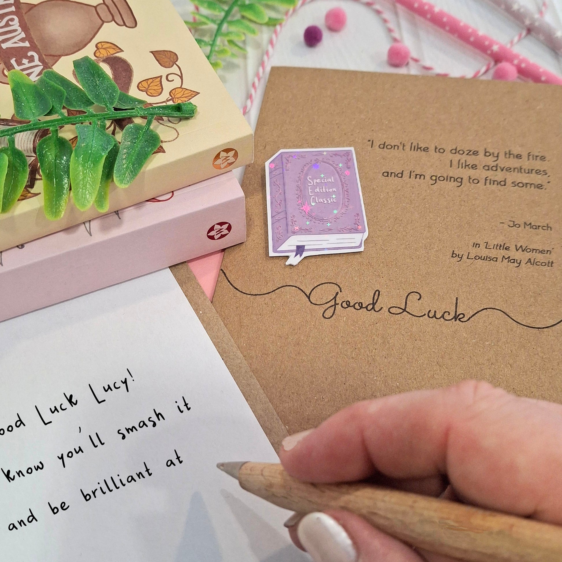 A close-up of a hand holding a wooden pen and writing a good luck message inside a card. The card features a raised holographic embellishment of a purple special edition classic book, the words Good Luck, and a quote from Little Women by Louisa May Alcott.