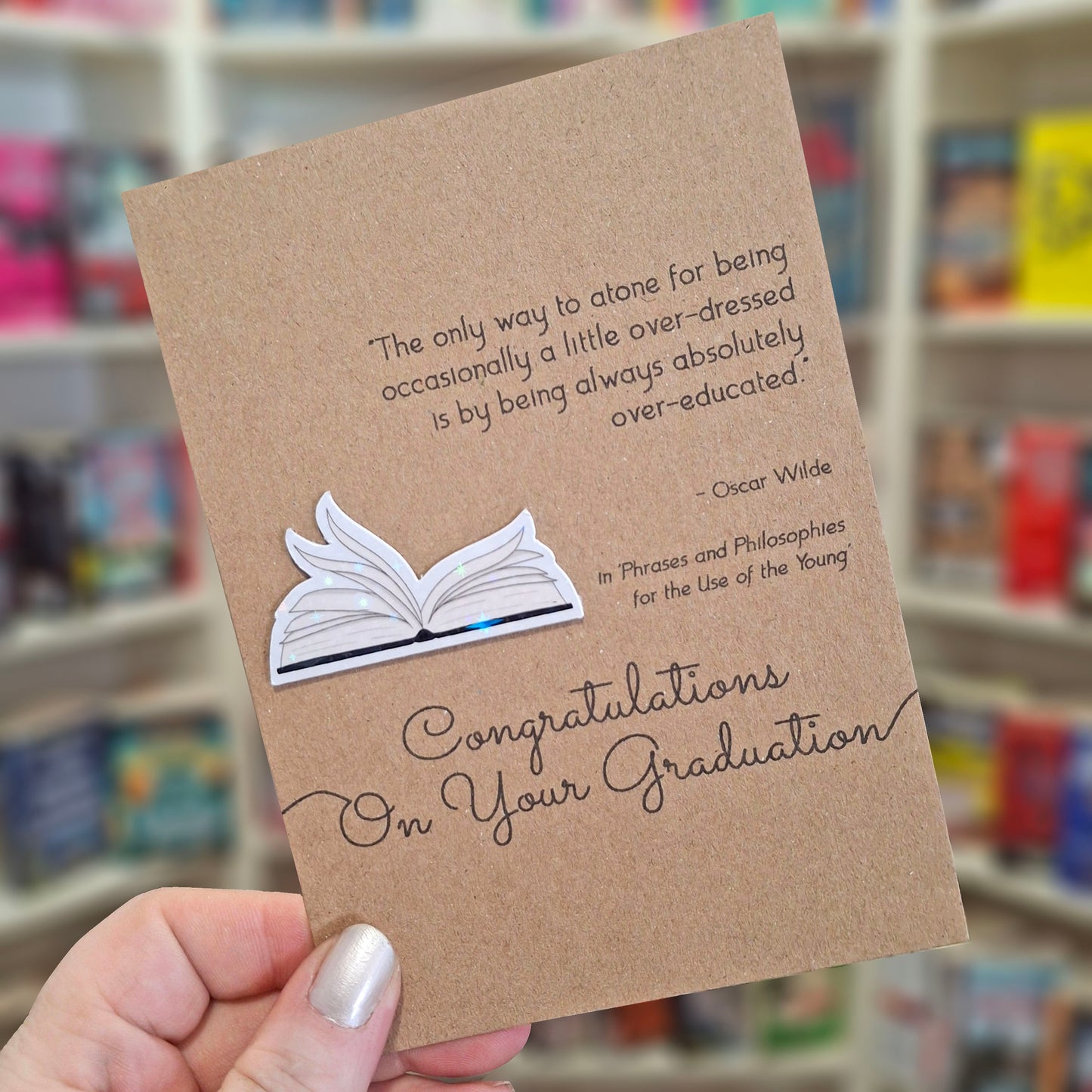 A hand holds a brown kraft congratulations on your graduation card featuring a raised holographic embellishment of an open book with gently fanned pages, and a printed quote by Oscar Wilde. The background shows a blurred bookshop interior.