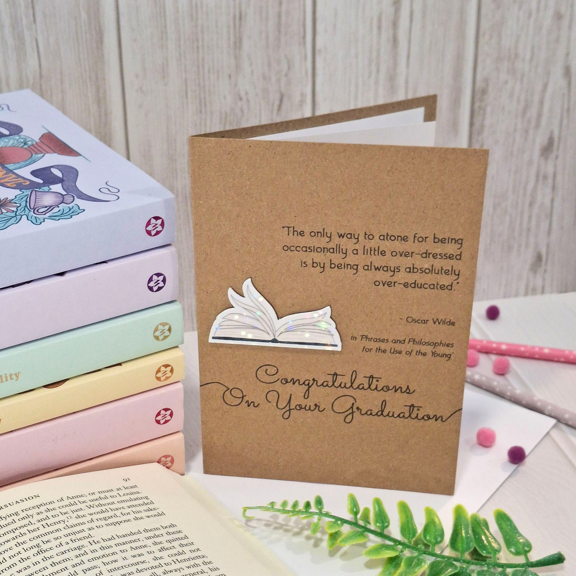 A brown kraft card styled on a light wood background with books and greenery. The card features a raised holographic embellishment of an open book with gently fanned pages, the words Congratulations on your Graduation, and a quote from Oscar Wilde.