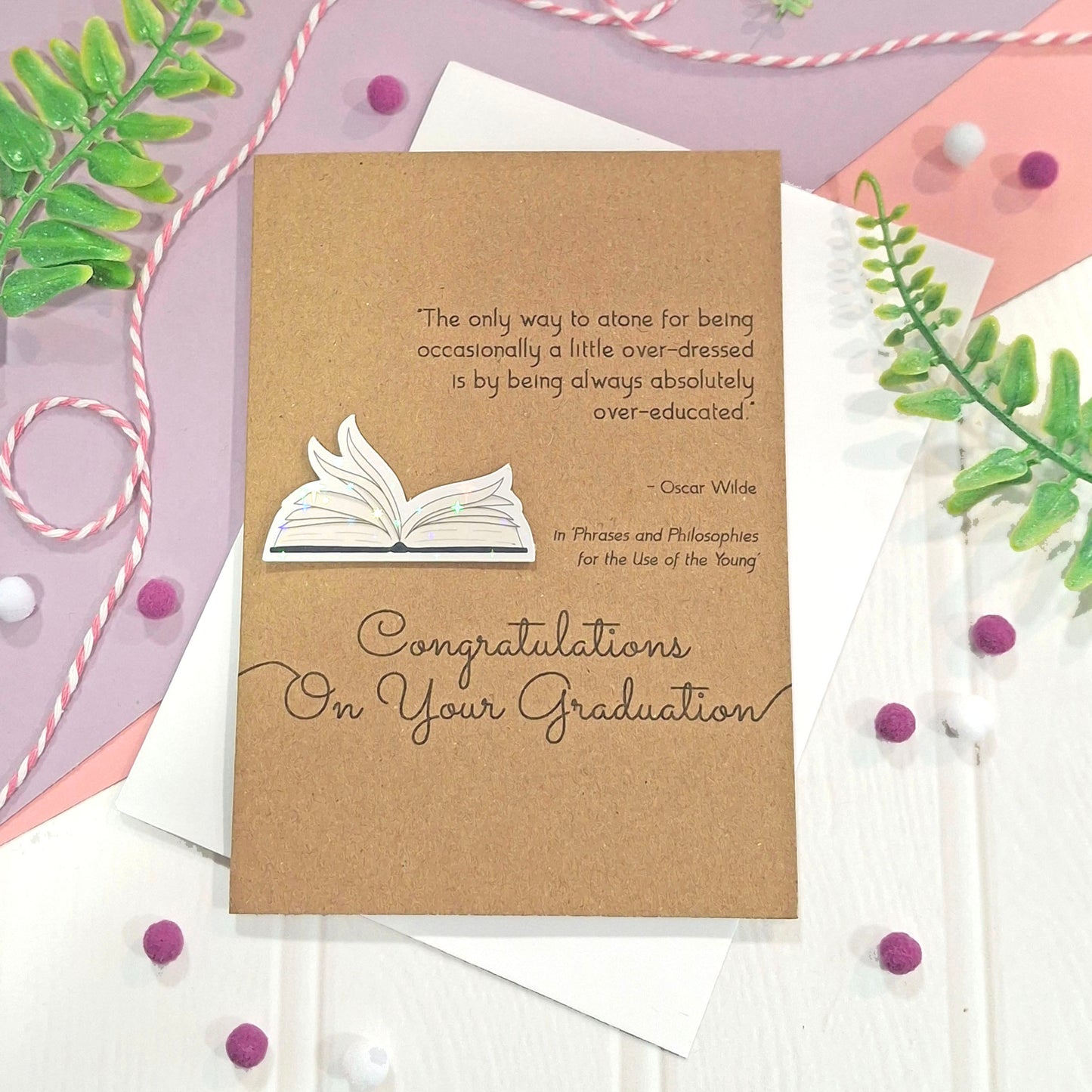A graduation card made from kraft card, featuring illustrated book pages in a raised, holographic embellishment, a quote by Oscar Wilde, and the words 'Congratulations On Your Graduation' in elegant script. The card is styled in a flat-lay arrangement with pastel paper, greenery, and small pom-poms.