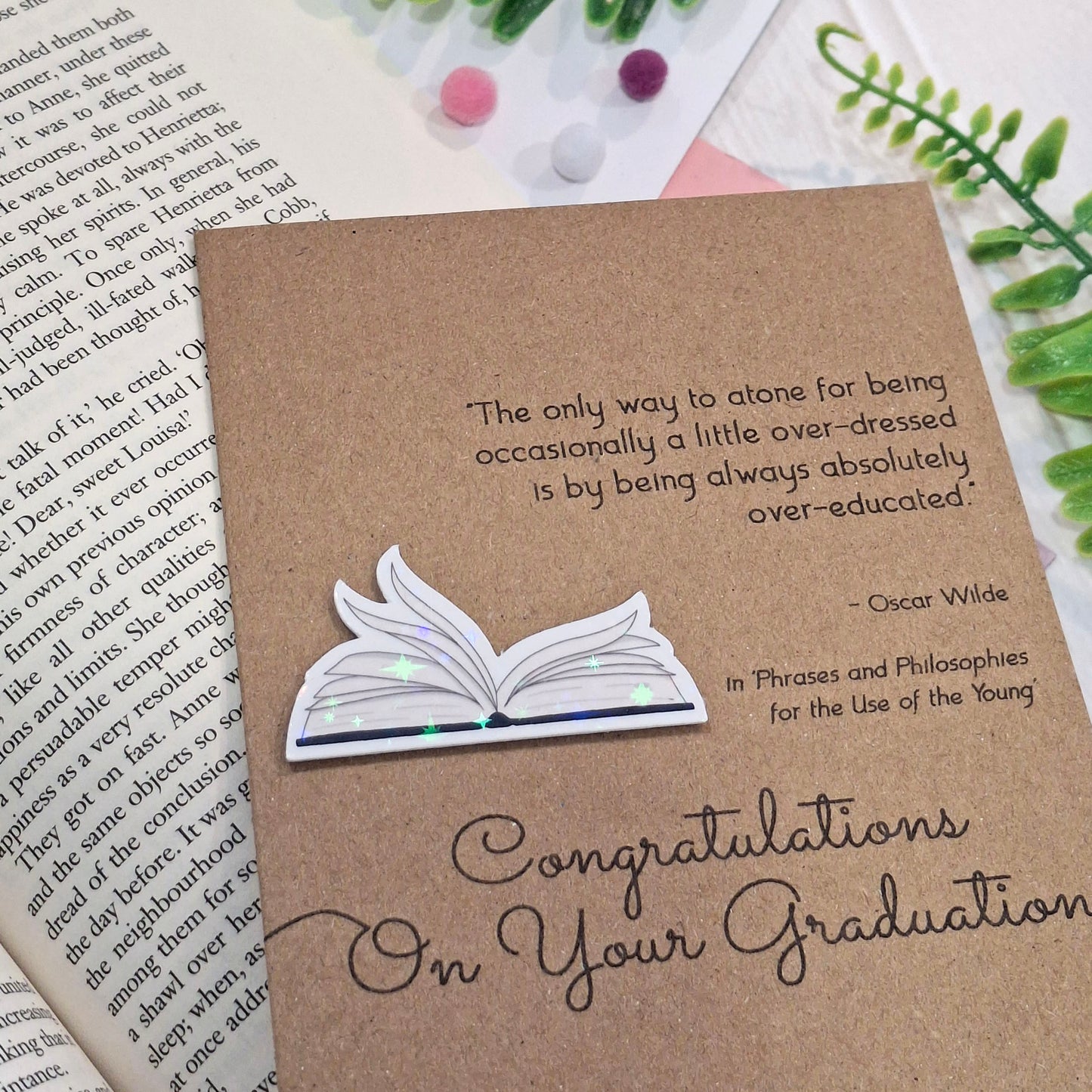 A brown kraft card styled on top of an open book with decorative pom-poms and greenery. The card features a raised holographic embellishment of an open book with gently fanned pages, the words Congratulations on your Graduation, and a quote from Oscar Wilde.