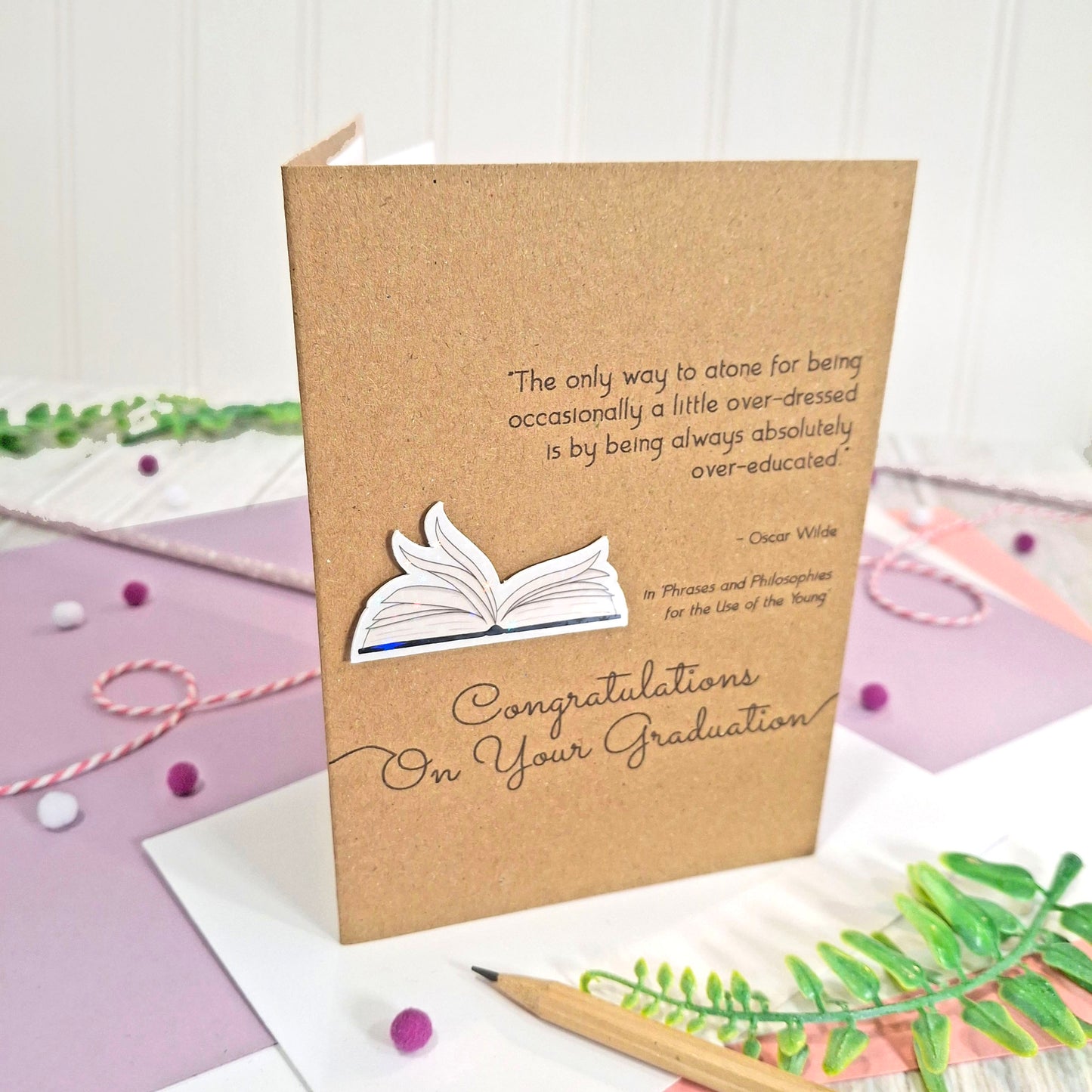 A handmade graduation card made from brown kraft card with a printed quote from Oscar Wilde, illustrated book pages in a raised, holographic embellishment, and 'Congratulations On Your Graduation' in black script. The card is styled on a white surface with a pencil, green leaves, and decorative threads.