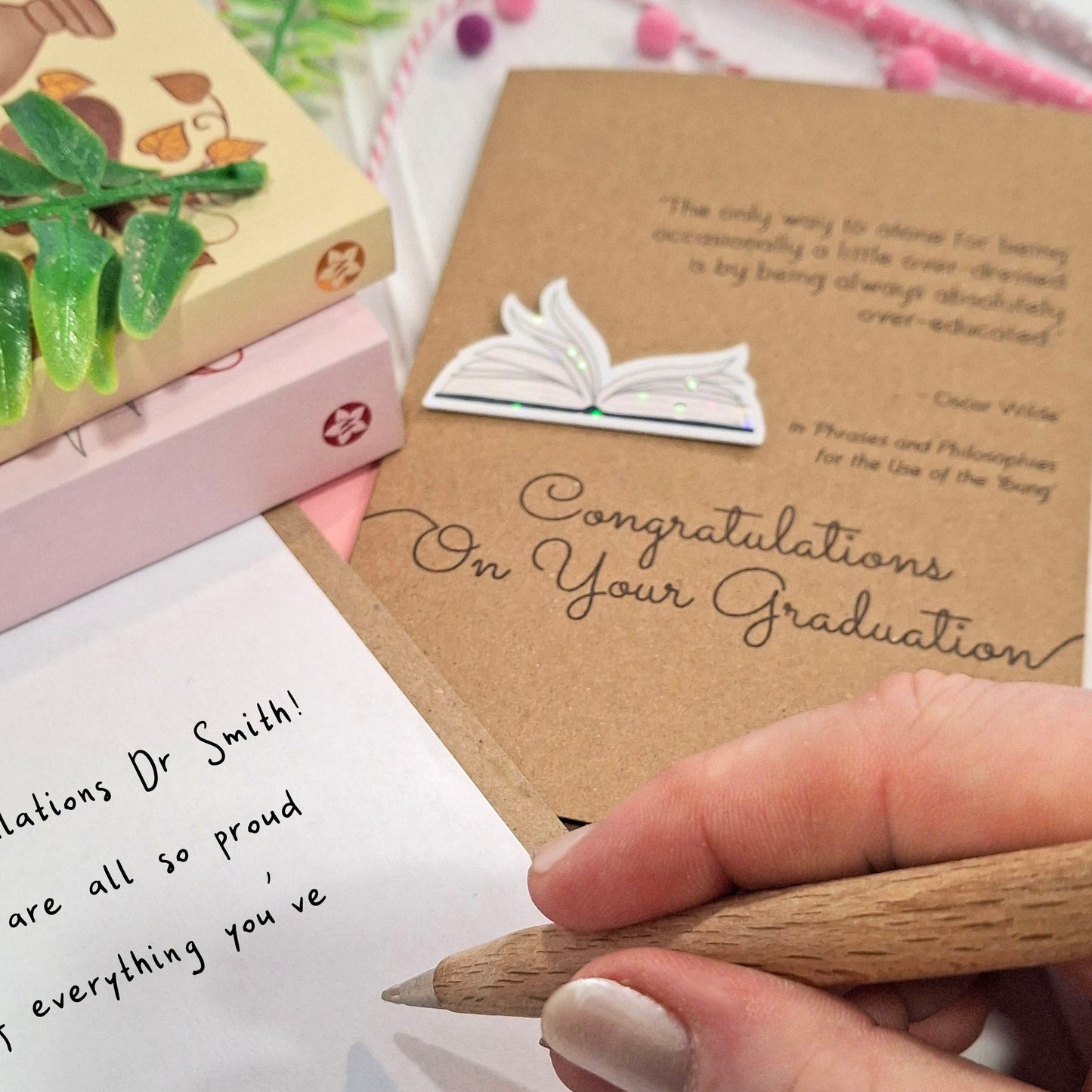 A close up of a hand holding a wooden pen, and writing a congratulations message inside a card. The card features a raised holographic embellishment of an open book with gently fanned pages, the words Congratulations on your Graduation, and a quote from Oscar Wilde.
