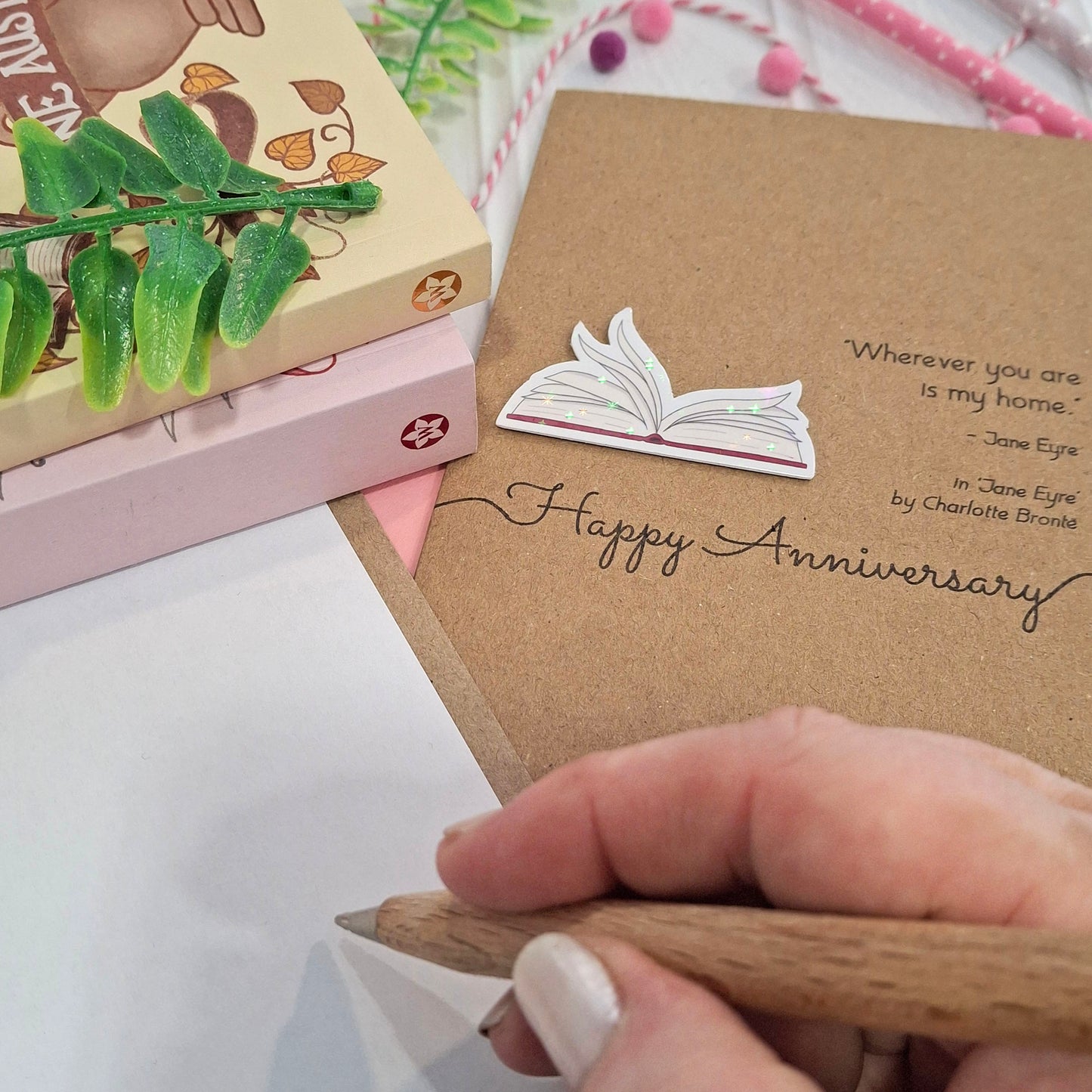 A close up of a hand holding a wooden pen, and writing inside a card. The card features a raised holographic embellishment of an open book with gently fanned pages, the words Happy Anniversary, and a quote from the book Jane Eyre by Charlotte Bronte.