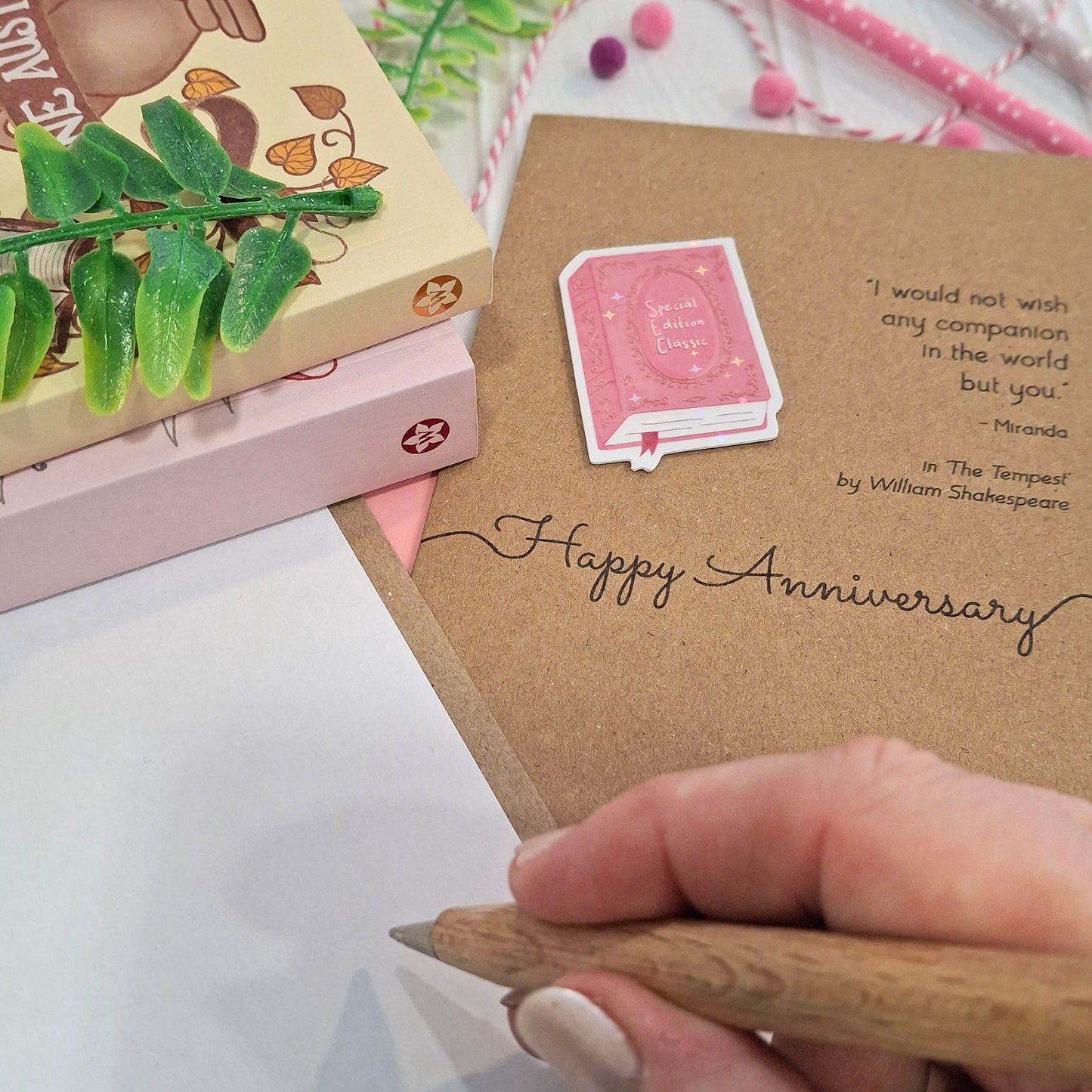 A close-up of a hand holding a wooden pen and writing inside a brown kraft anniversary card. The card features a raised holographic embellishment of a pink special edition classic book, the words Happy Anniversary, and a quote from The Tempest by William Shakespeare.