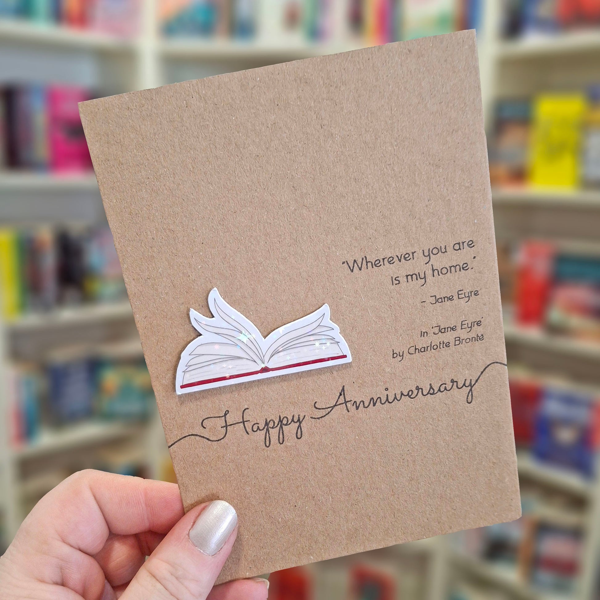A hand holds a brown kraft happy anniversary card featuring a raised holographic embellishment of an open book with gently fanned pages, and a printed quote by Charlotte Bronte. The background shows a blurred bookshop interior.