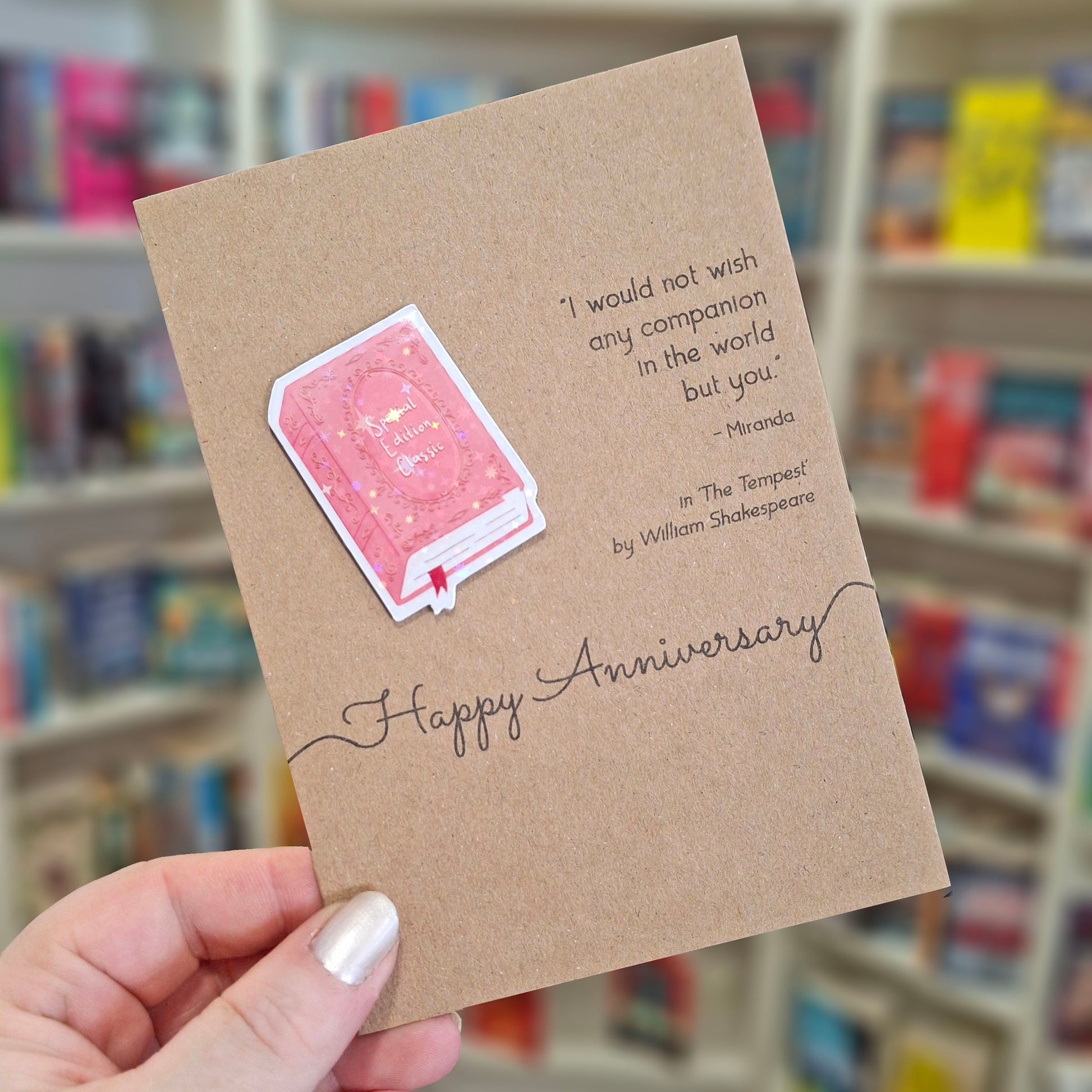 A hand holds a brown kraft happy anniversary card featuring a raised holographic embellishment of a pink special edition classic book, and a printed quote by William Shakespeare. The background shows a blurred bookshop interior.