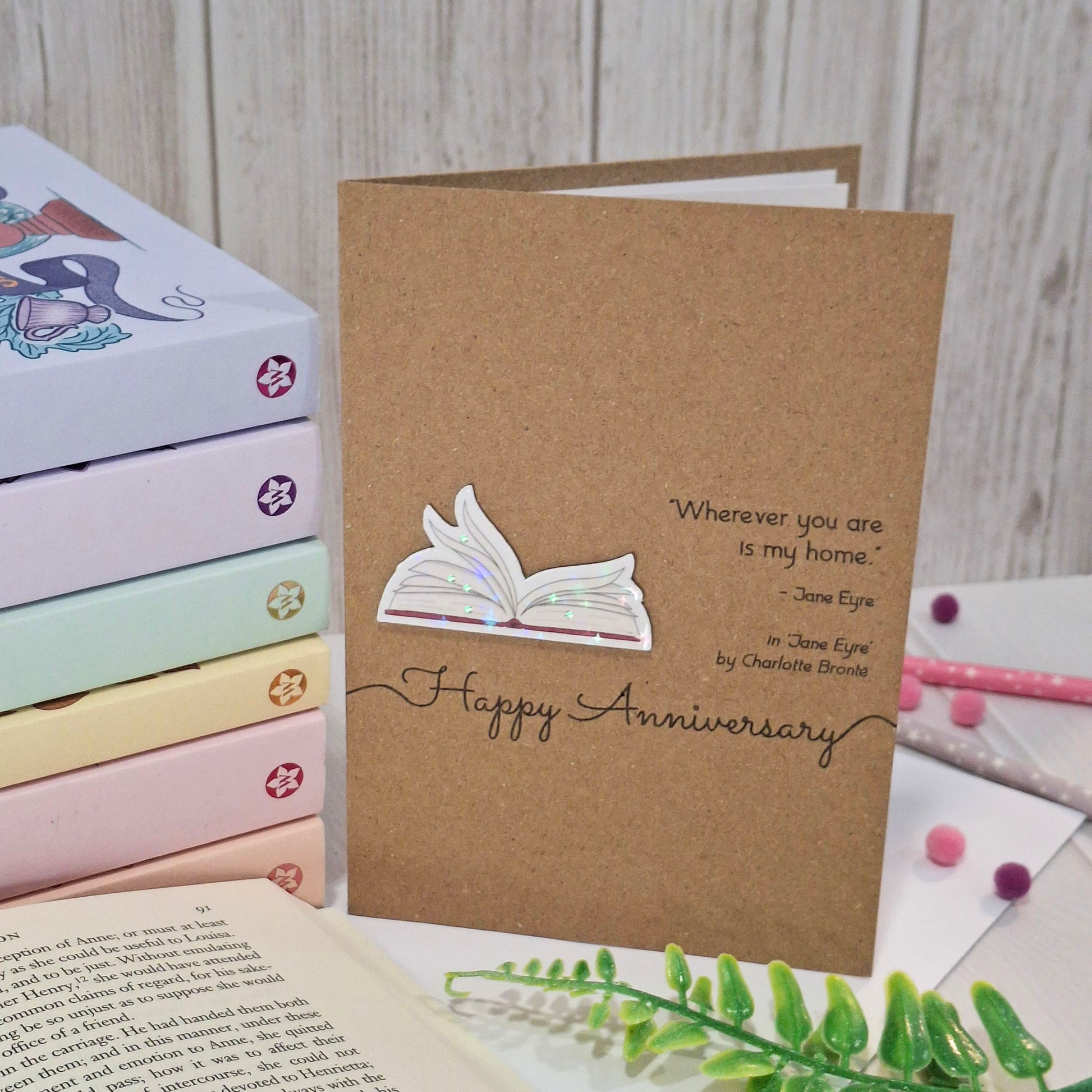 A handmade brown kraft anniversary card displayed on a tabletop with other books and decorative items. The card features a raised holographic embellishment of an open book with gently fanned pages, the words Happy Anniversary, and a quote from the book Jane Eyre by Charlotte Bronte.