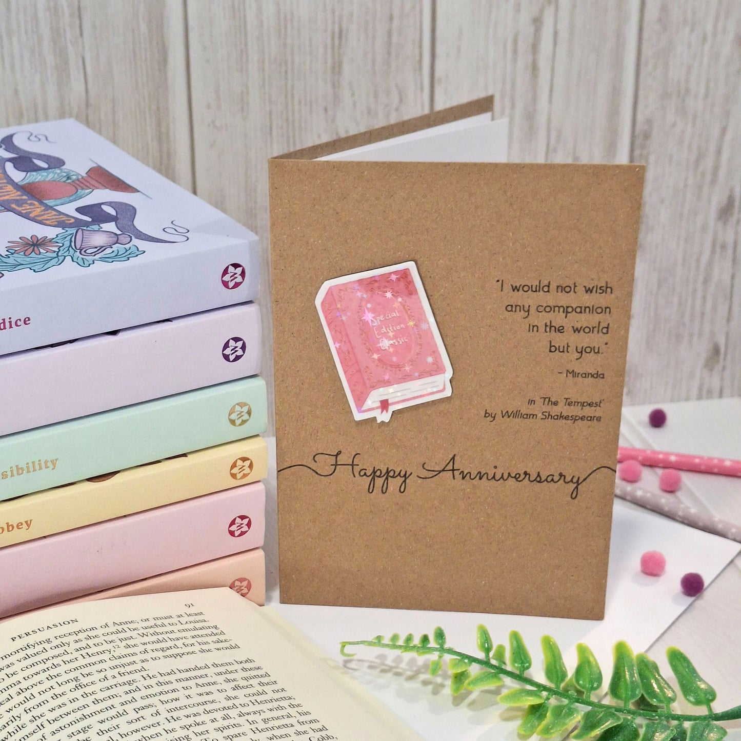 A kraft anniversary card styled on a light wood background with books, a book page, and decorative greenery. The card features a raised holographic embellishment of a pink special edition classic book, the words Happy Anniversary, and a quote from The Tempest by William Shakespeare.