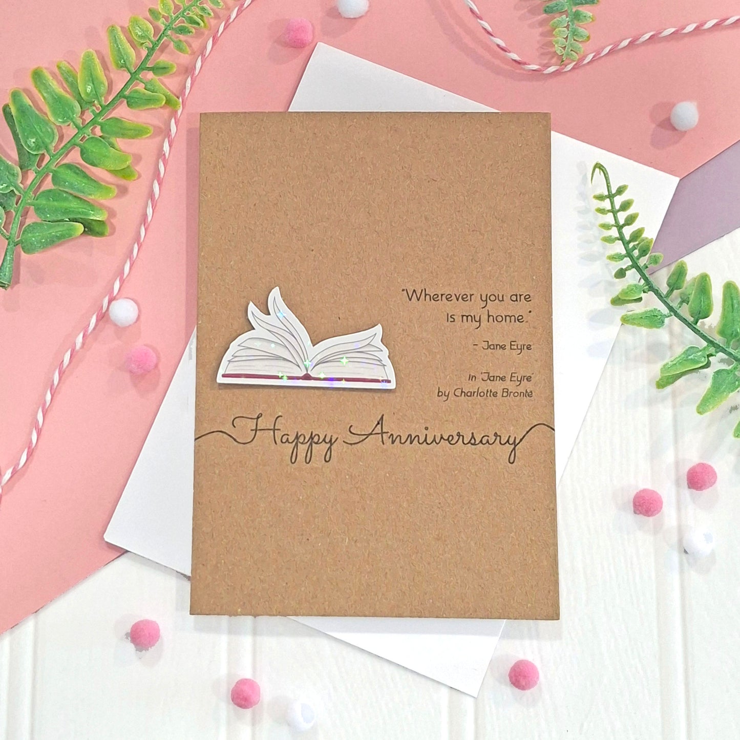 A card that reads 'Happy Anniversary' made from brown kraft card, featuring illustrated book pages in a raised, holographic embellishment, and printed quote from the book Jane Eyre by Charlotte Bronte, styled on white and pink paper with decorative green leaves and pink pom-poms.