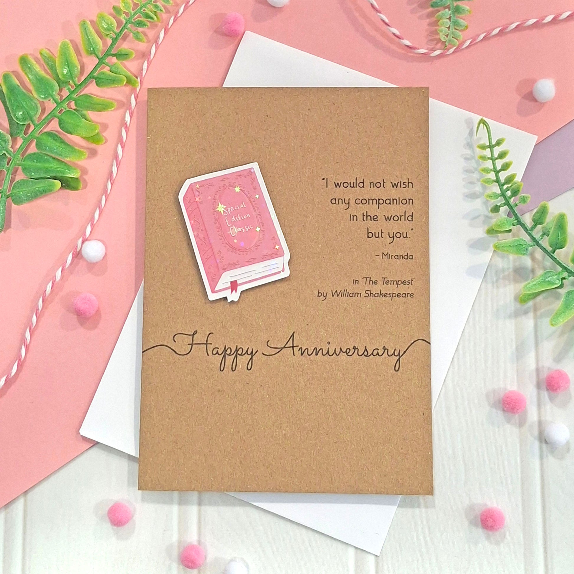 A brown kraft greeting card with a raised, holographic, illustrated story book embellishment in pink and a quote from William Shakespeare's The Tempest. The card reads 'Happy Anniversary' and is styled on top of white envelopes with pink and white pom-poms and green leaves.