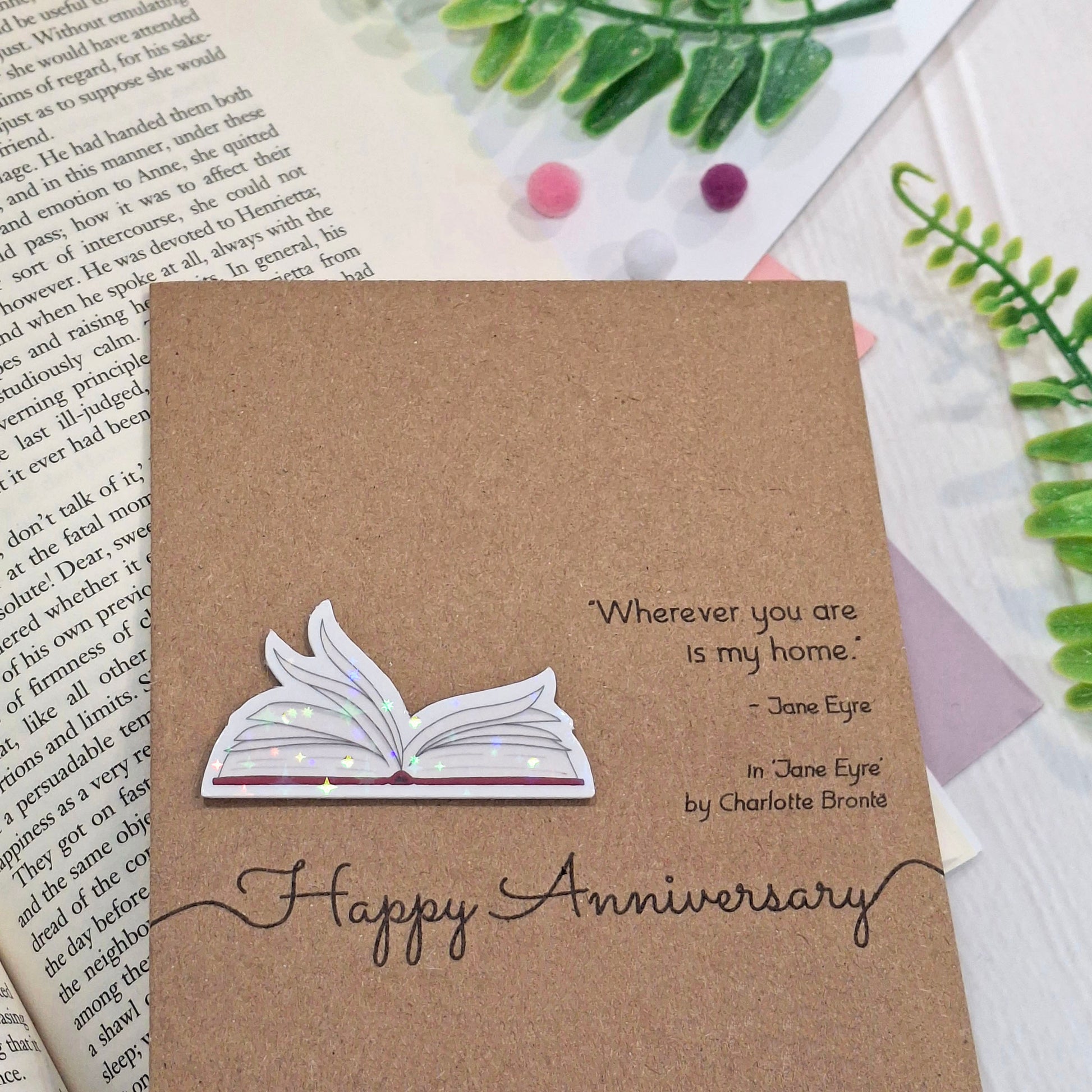A handmade brown kraft anniversary card styled on top of an open book with decorative green leaves and small pink and purple beads. The card features a raised holographic embellishment of an open book with gently fanned pages, the words Happy Anniversary, and a quote from the book Jane Eyre by Charlotte Bronte.