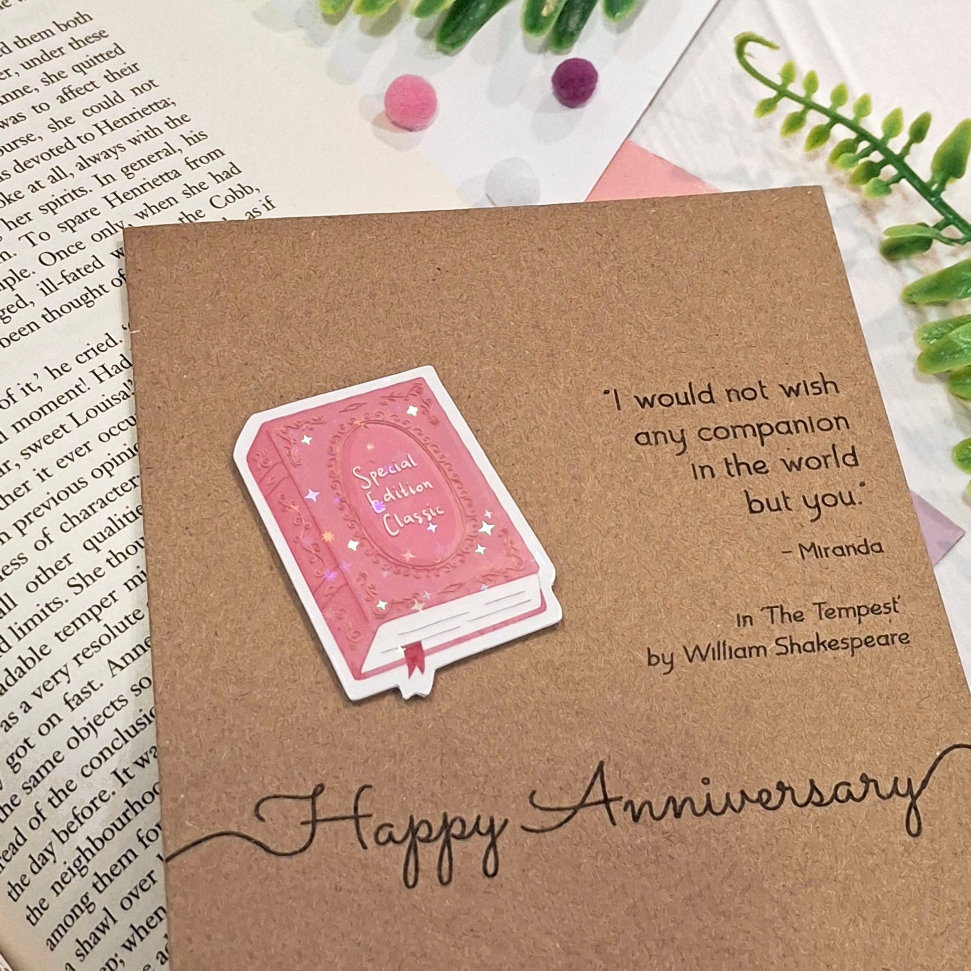 A brown kraft anniversary card styled on top of an open book with decorative greenery and pom-poms. The card features a raised holographic embellishment of a pink special edition classic book, the words Happy Anniversary, and a quote from The Tempest by William Shakespeare.