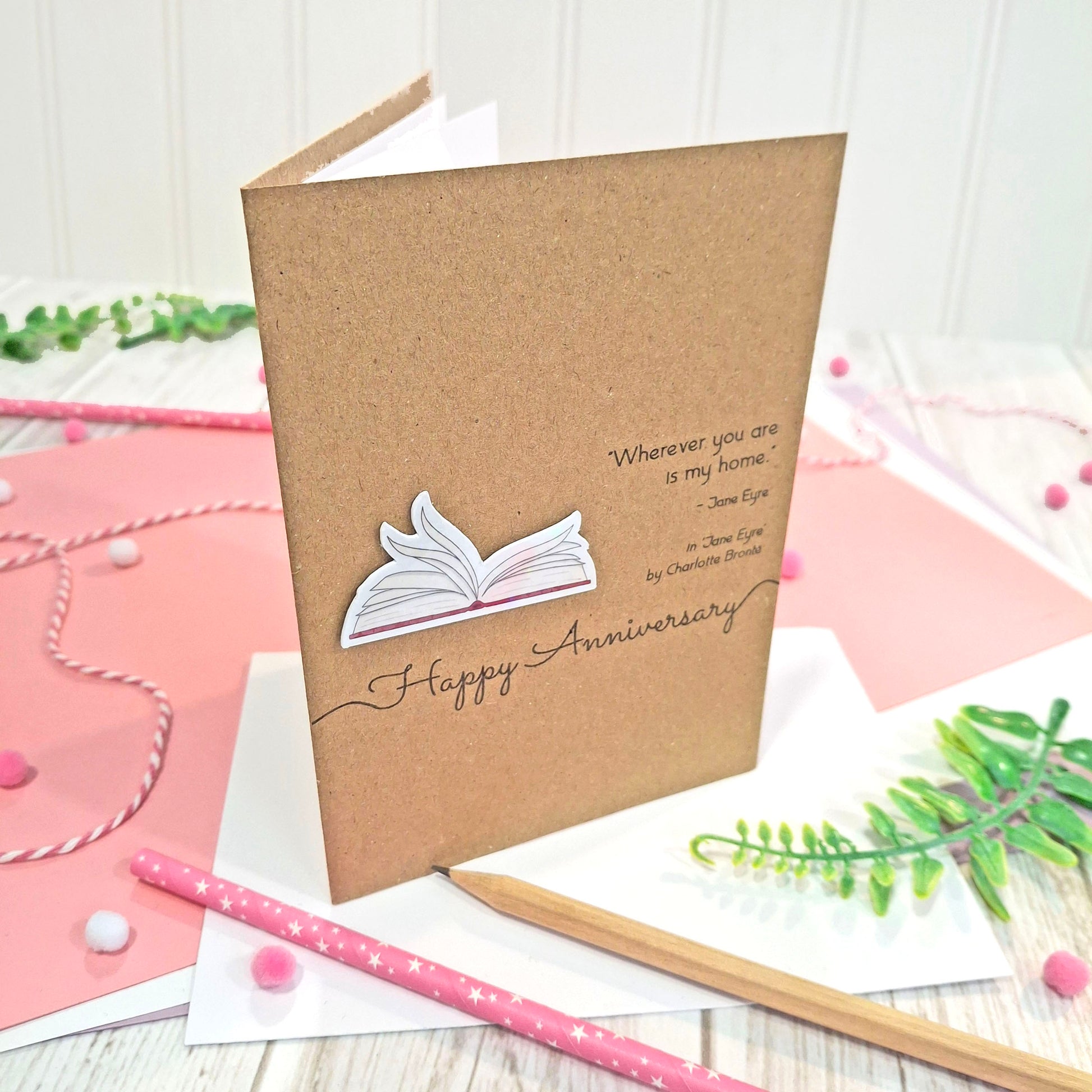 A brown kraft brown happy anniversary card with illustrated book pages in a raised, holographic embellishment and a quote from Jane Eyre by Charlotte Bronte.  The card reads 'Happy Anniversary' in elegant script, and is styled on a white wood surface with pink stationery and greenery.