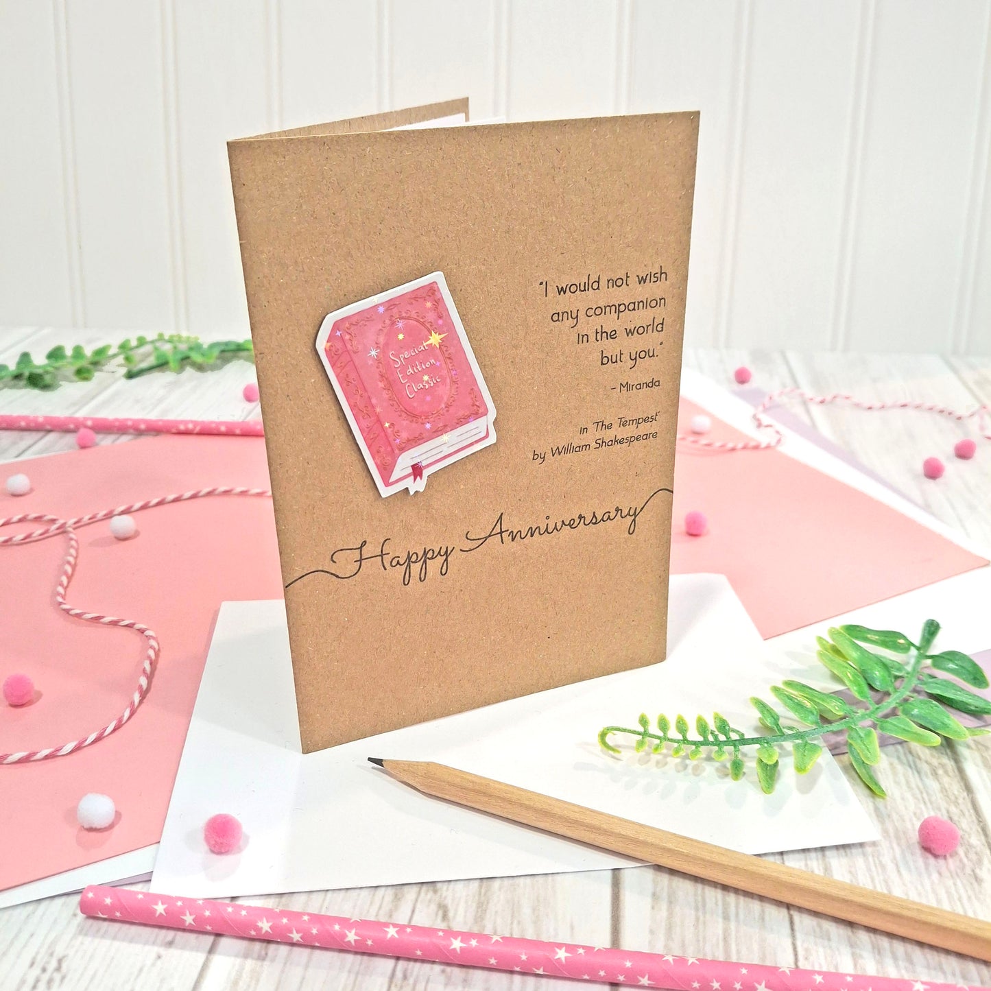 A handmade brown kraft anniversary card with  a raised, holographic, illustrated book embellishment in pink, the words 'Happy Anniversary' and a quote from The Tempest by William Shakespeare, all displayed on a styled flat-lay surface with pink and white paper, a wooden pencil, and greenery.