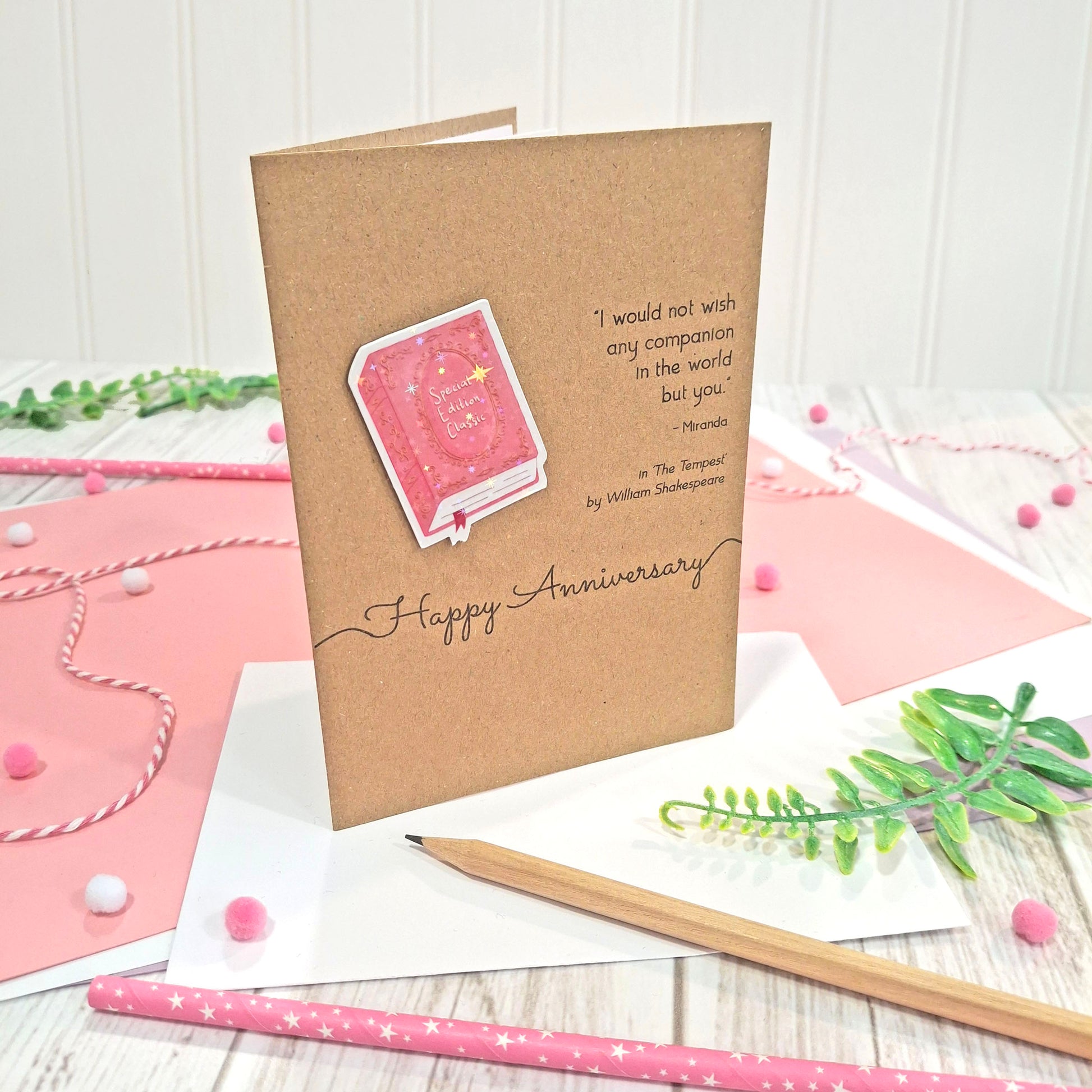 A handmade brown kraft anniversary card with  a raised, holographic, illustrated book embellishment in pink, the words 'Happy Anniversary' and a quote from The Tempest by William Shakespeare, all displayed on a styled flat-lay surface with pink and white paper, a wooden pencil, and greenery.